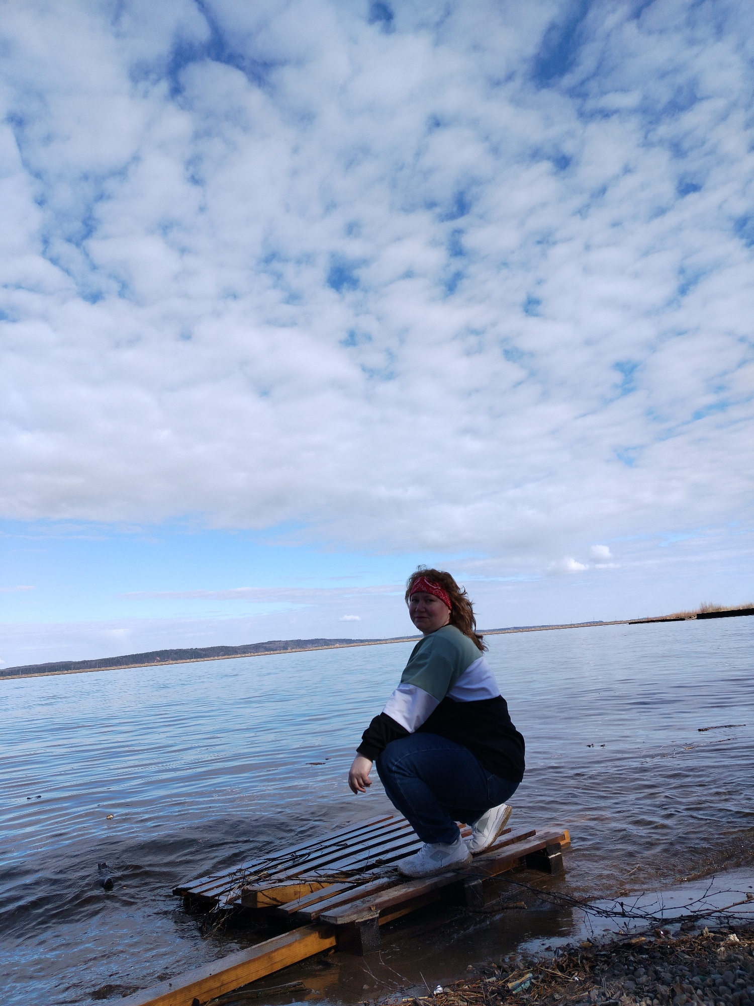 A person with reddish-brown hair and wearing a red bandana, a green, white, and black sweatshirt, blue jeans, and white sneakers is crouching on a wooden pallet that is partially submerged in water near the shoreline of a body of water. The water extends to the horizon, where a strip of land is visible in the distance. The sky is predominantly blue with scattered white clouds. The shoreline consists of sand, pebbles, and some debris. The location is Chaikovsky, Russia.