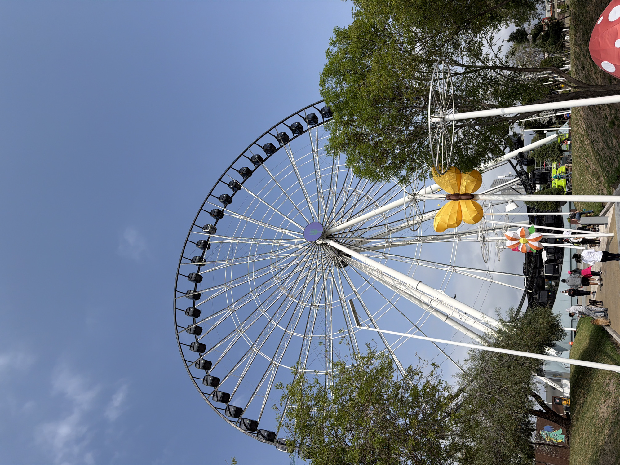 The image shows the Puebla Star Ferris wheel in Puebla, Mexico. It is a clear day, and the Ferris wheel dominates the left side of the image. The wheel is white with black gondolas and a blue center. A large yellow butterfly decoration is visible on a white structure near the Ferris wheel. Trees partially obscure parts of the wheel and the structure. On the right side of the image, a path with several people walking along it can be seen. A mushroom-shaped decoration is also visible.