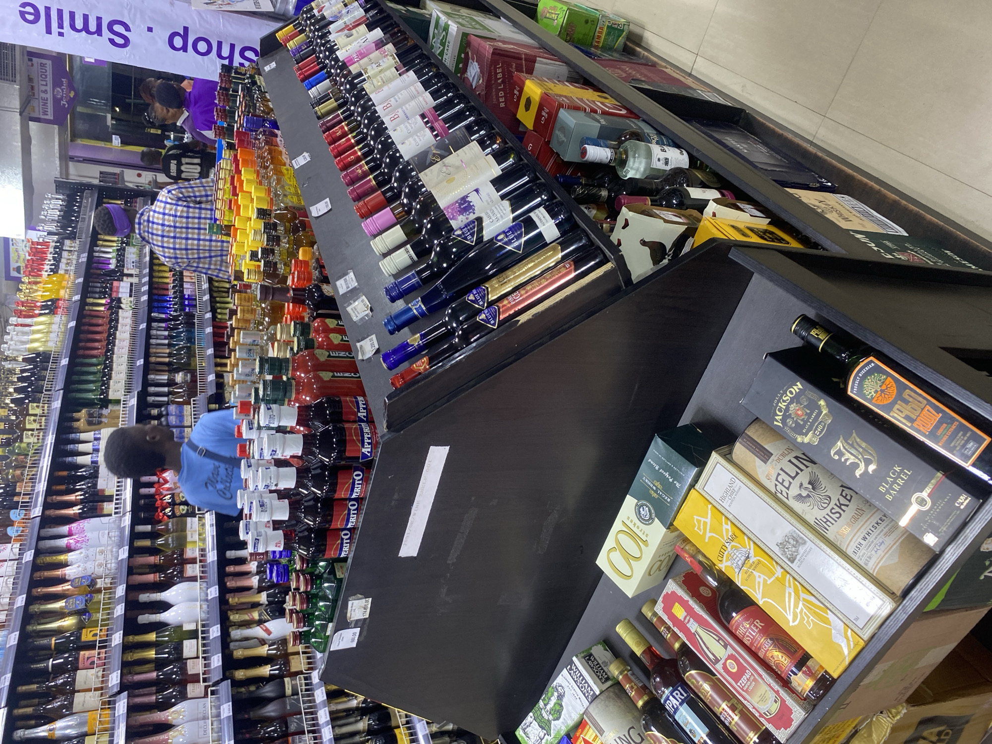 The image depicts the interior of a liquor store, likely in Lagos, Nigeria, recognizable by the product labels and signage.
A man wearing a blue shirt stands in front of shelves stocked with a variety of wines and liquors. Shelves displaying wine and spirits dominate the scene. Bottles of wines and spirits are arranged on the shelves and on a central display unit. A second person, also possibly a customer, is visible in the background.
The image is an amateur photograph taken indoors.