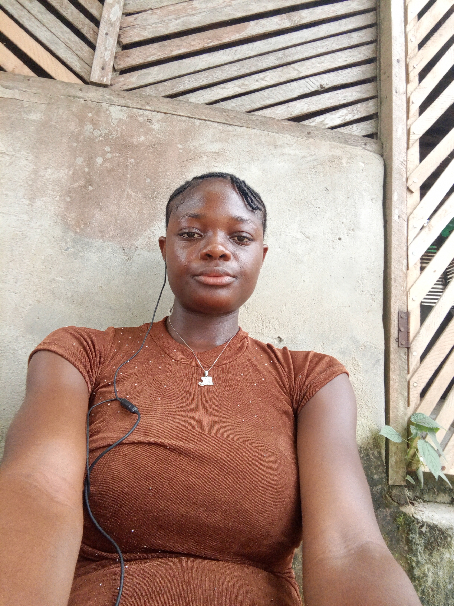 A young woman is the primary subject of the image. She has dark skin, short braided hair, and is wearing a brown shirt with small, shiny embellishments. A black wired earphone is draped around her neck. She is wearing a silver necklace with a pendant.
She appears to be outdoors, leaning against a wall. The wall is made of concrete, and there is a wooden structure behind and to the right of the wall. Green plants are visible near the base of the wooden structure. The location is Mbribit, Nigeria.