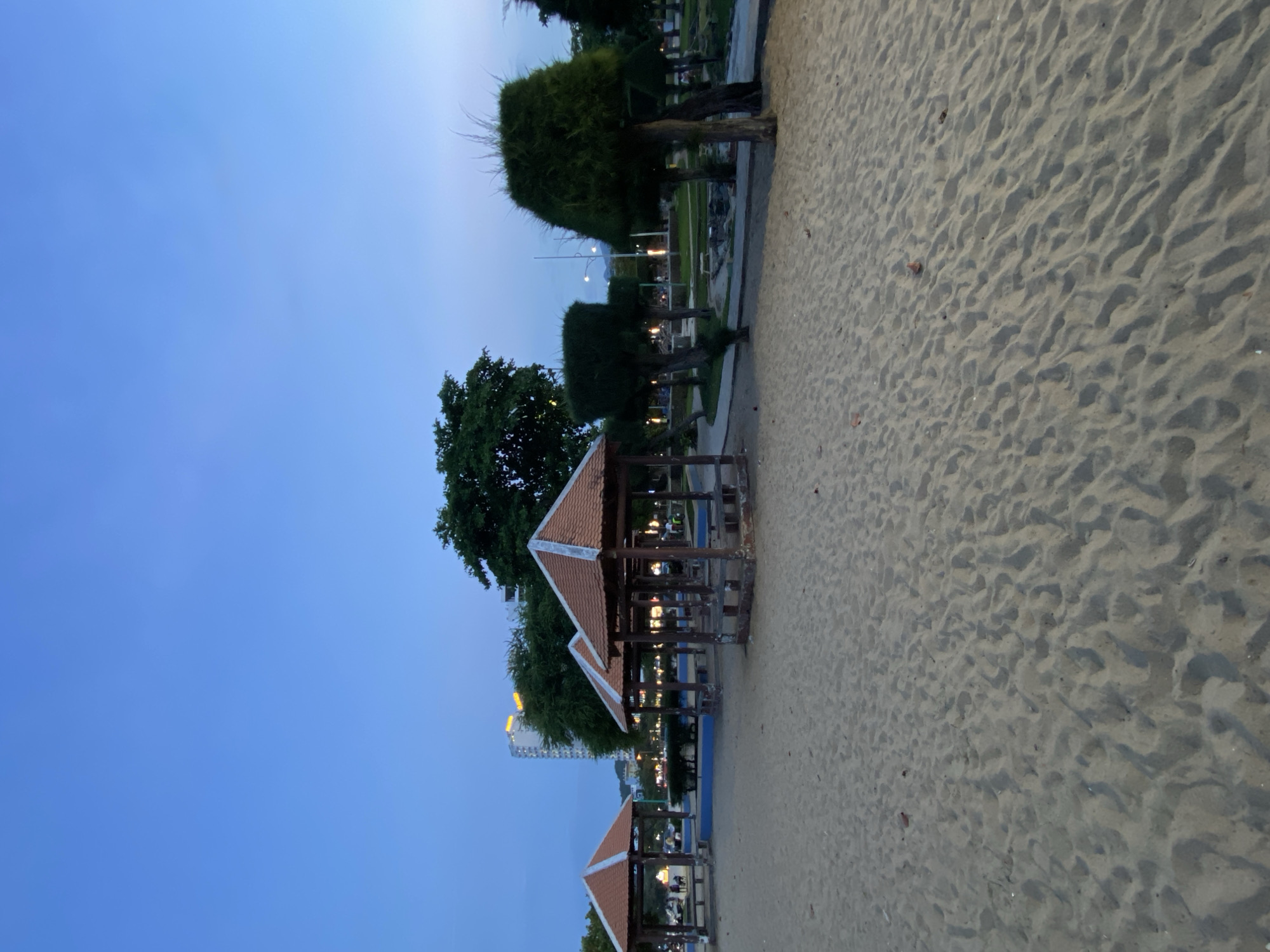 The image depicts a sandy beach area with several open-air shelters featuring terracotta-tiled roofs and wooden posts. These shelters appear to be gazebos, each containing built-in benches. In the background, there is a lush green park with manicured trees, some of which are distinctively shaped. Beyond the park, city buildings, including a tall, light-colored high-rise with illuminated windows, are visible against a twilight sky. The overall scene suggests a public recreational space in Nha Trang, Vietnam, likely near a beach or promenade. The lighting indicates it is dusk or early evening, with artificial lights beginning to illuminate the park and city. There are a few indistinct figures visible in the distance, but no prominent individuals or specific interactions are depicted. The foreground is dominated by the texture of the sand, showing ripples and footprints.