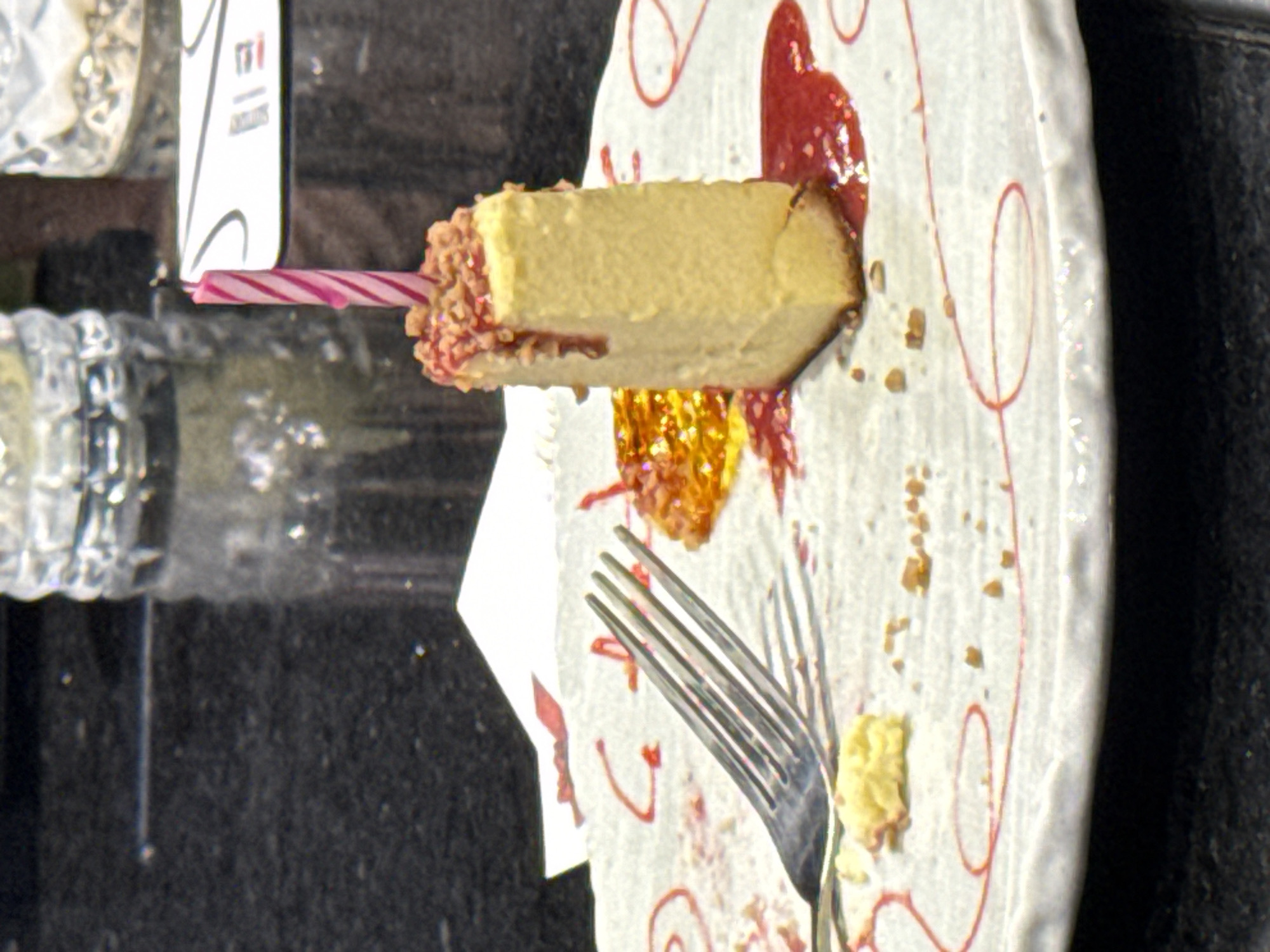 This close-up image captures a slice of cheesecake resting on a white plate, placed on a dark, textured surface, likely a table. A pink and white striped birthday candle is inserted into one end of the cheesecake, suggesting a celebration. The cheesecake is a light yellow color with a slightly browned crust on its base and is topped with reddish, crushed nuts or candy on the end closest to the candle.
The white plate is adorned with delicate red squiggly lines and features dollops of a vibrant red sauce, possibly fruit jam, some of which sparkle with golden glitter. One section of the red sauce appears somewhat heart-shaped. Scattered cheesecake crumbs on the plate indicate that the dessert has been partially eaten. A silver-colored fork with four tines rests on the plate, positioned to the right of the cheesecake.
In the background, a white piece of paper or card is partially visible, displaying the letters "TFI" and some smaller, illegible text, likely part of a restaurant or brand logo. To the left, a section of a clear, possibly faceted glass or plastic container can be seen. The birthday candle is unlit. The image depicts a moment of enjoying dessert, likely in a casual dining setting.