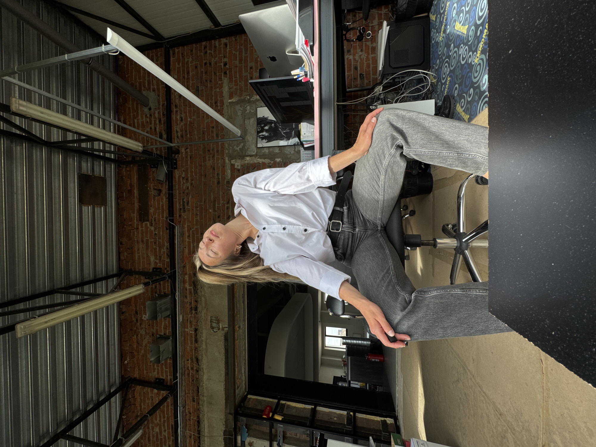 A young Caucasian woman is seated in a swivel chair in what appears to be a loft-style office space. She is wearing a white button-down shirt and grey jeans. Her eyes are closed and her head is tilted back, suggesting she is resting or stretching. The office has exposed brick walls, a metal ceiling with exposed beams and lighting fixtures, and a desk with computers and office supplies. The environment appears somewhat unfinished or industrial, with some construction elements visible. There is no identifiable connection to Chaikovsky, Russia.