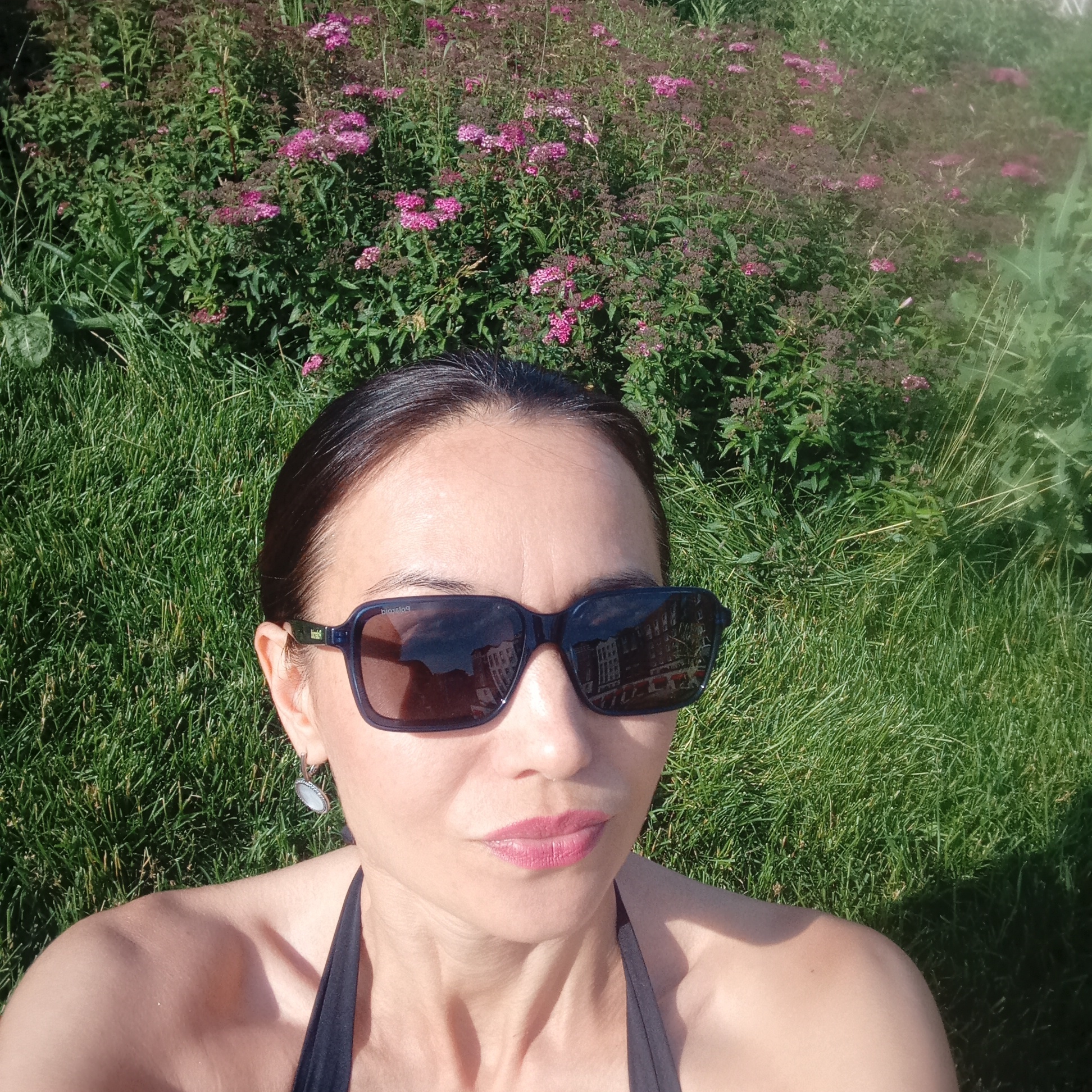 A close-up, high-angle shot shows a woman lying on a patch of green grass in front of a dense bush of pink and purple flowers. The woman is wearing dark sunglasses, a black halter top, and a silver earring. Her lips are slightly parted, and she is wearing pink lipstick. The sunglasses reflect buildings. The scene appears to be outdoors on a sunny day. The location is identified as Pervomaysky, Russia.