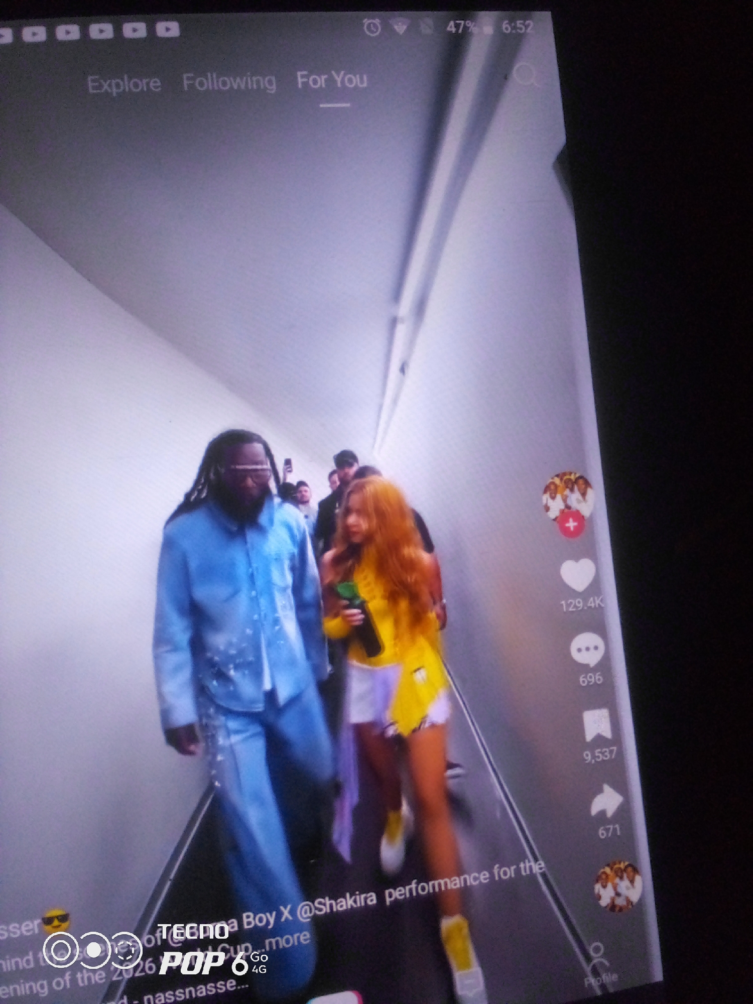 The image depicts a screen recording of a social media video, likely TikTok, showing two individuals walking down a brightly lit hallway. The person on the left is a Black male with dreadlocks, wearing a light blue patterned shirt and matching pants. He has on glasses. The person on the right is a female with reddish-blonde hair, wearing a yellow top and white shorts. She is holding a green bottle. Several other figures are visible in the background, also walking.
On the right side of the screen, social media engagement icons are visible: a profile picture with a '+' icon, a heart icon with the number 129.4K, a speech bubble icon with the number 696, a bookmark icon with the number 9,537, and a share icon with the number 671.
At the top of the screen, navigation options read "Explore," "Following," and "For You," with "For You" highlighted. The time displayed is 6:52 and the battery level is 47%.
In the bottom left corner, there is text that appears to be part of the video's caption or overlay. It includes the word "TECNO" and "POP 8" along with "Go 4G," suggesting this is a Tecno Pop 8 phone. Other visible text mentions "Boy X @Shakira performance" and "nass...".
The environment is an indoor hallway with white walls and a white ceiling. The floor appears to be made of a dark material.
There is no information in the image that specifies the location as Howayi, Nigeria.