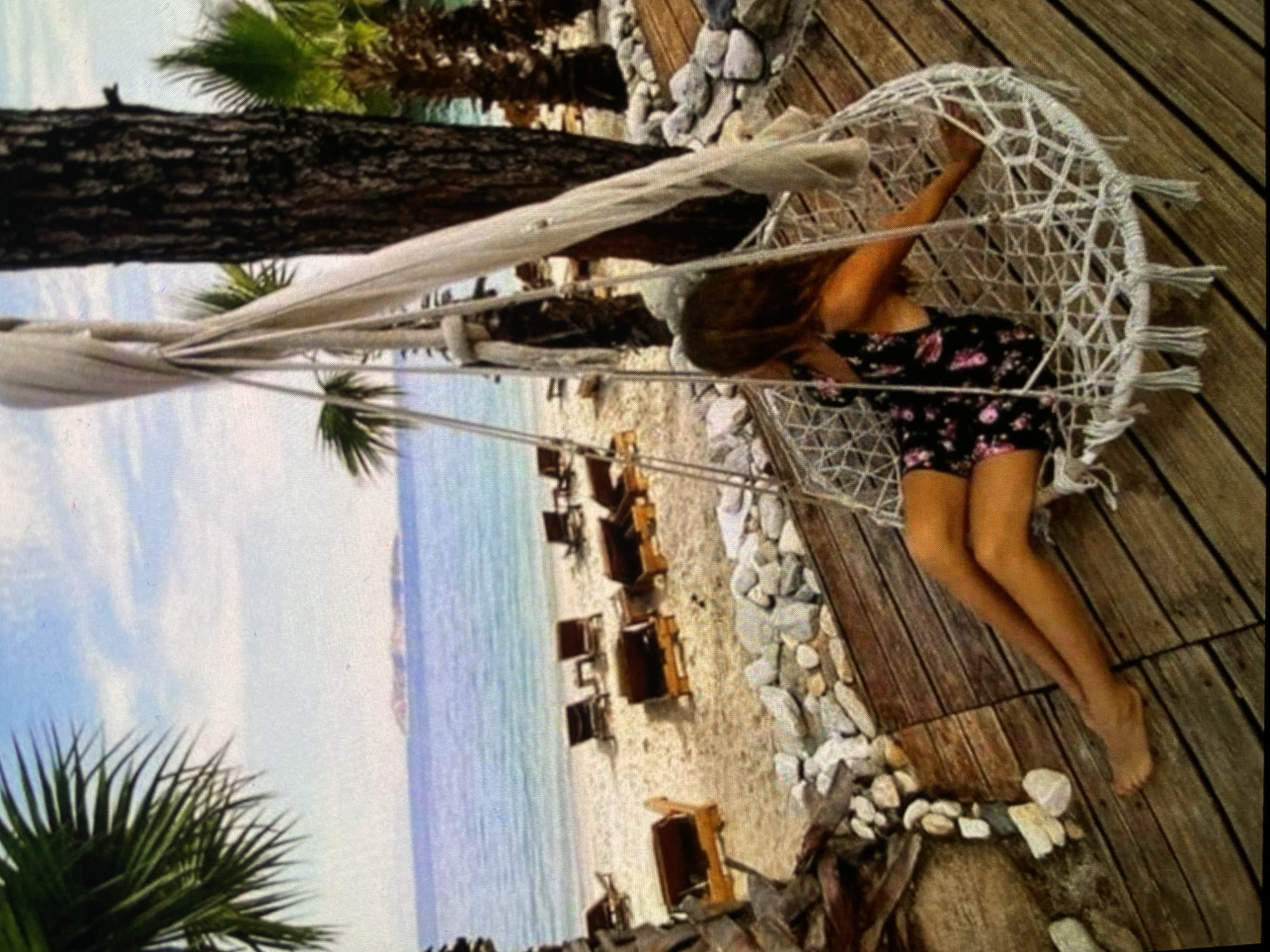 The image, rotated 90 degrees counter-clockwise, displays a relaxing daytime scene at what appears to be a coastal resort. The central subject is a young woman, seen from the back, with long brown hair, wearing a dark floral-print dress or romper. She is comfortably nestled inside a white, woven, spherical hanging chair. Her bare legs are extended and visible against the wooden deck where the chair is situated.
The setting includes a wooden deck in the foreground and right, partially covered with scattered light-colored rocks. To the left, a thick tree trunk horizontally crosses the upper mid-ground, with vibrant green palm fronds visible behind it and in the lower left corner. Beyond the rocky edge, a sandy beach is visible, lined with several empty wooden beach chairs. The calm, light blue sea stretches out to the horizon under a bright, partly cloudy sky, indicating sunny weather. The atmosphere is peaceful and leisurely. No text is visible in the image. The location context provided is Bankya, Bulgaria.