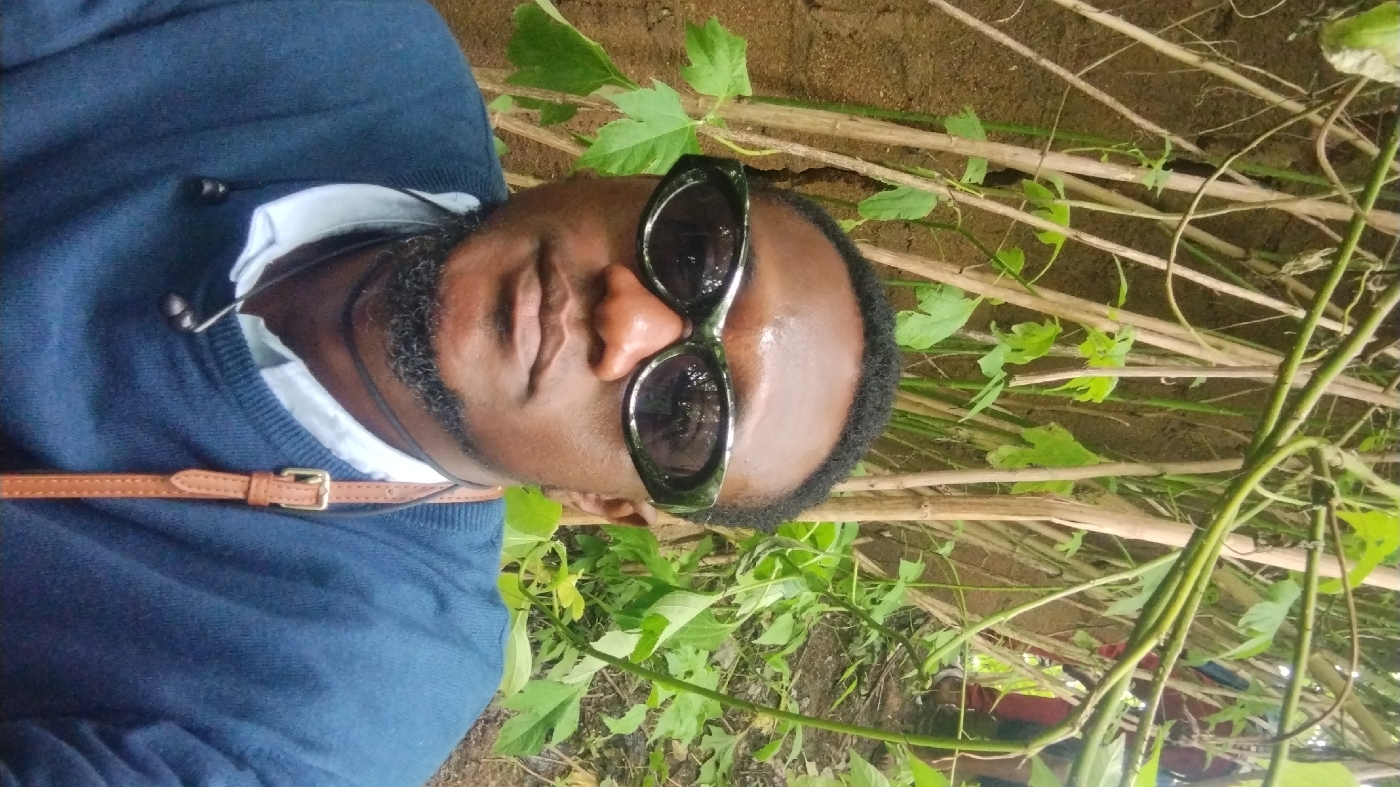 The image is a selfie taken outdoors, likely in a natural, overgrown setting. A dark-skinned man wearing sunglasses, a blue sweater over a light-colored shirt, and a brown strap across his chest is the main subject. The background consists of a wall of thin, dry stalks and green foliage, suggesting a dense, possibly wild or uncultivated area. There are hints of other people in the background, wearing red and blue, further suggesting a communal or outdoor activity. The lighting is natural and appears to be daylight. The image captures the man from the chest up, with the background slightly out of focus. There are no specific landmarks or indications of the city of Jos, Nigeria, visible in the image.