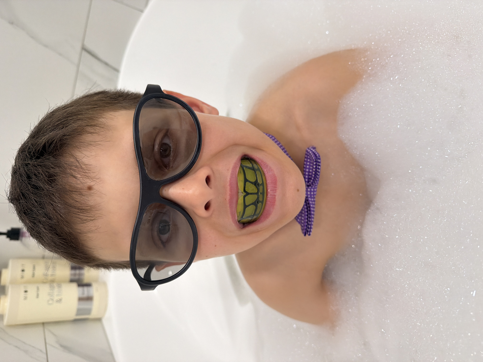 A boy is in a bathtub filled with white bubbles. He is wearing large, black-framed glasses and a purple polka-dotted bow tie. A green and black mouthguard is in his mouth. Two bottles of hair care products are visible on the side of the tub. The location is in a bathroom, potentially in Votkinsk, Russia.