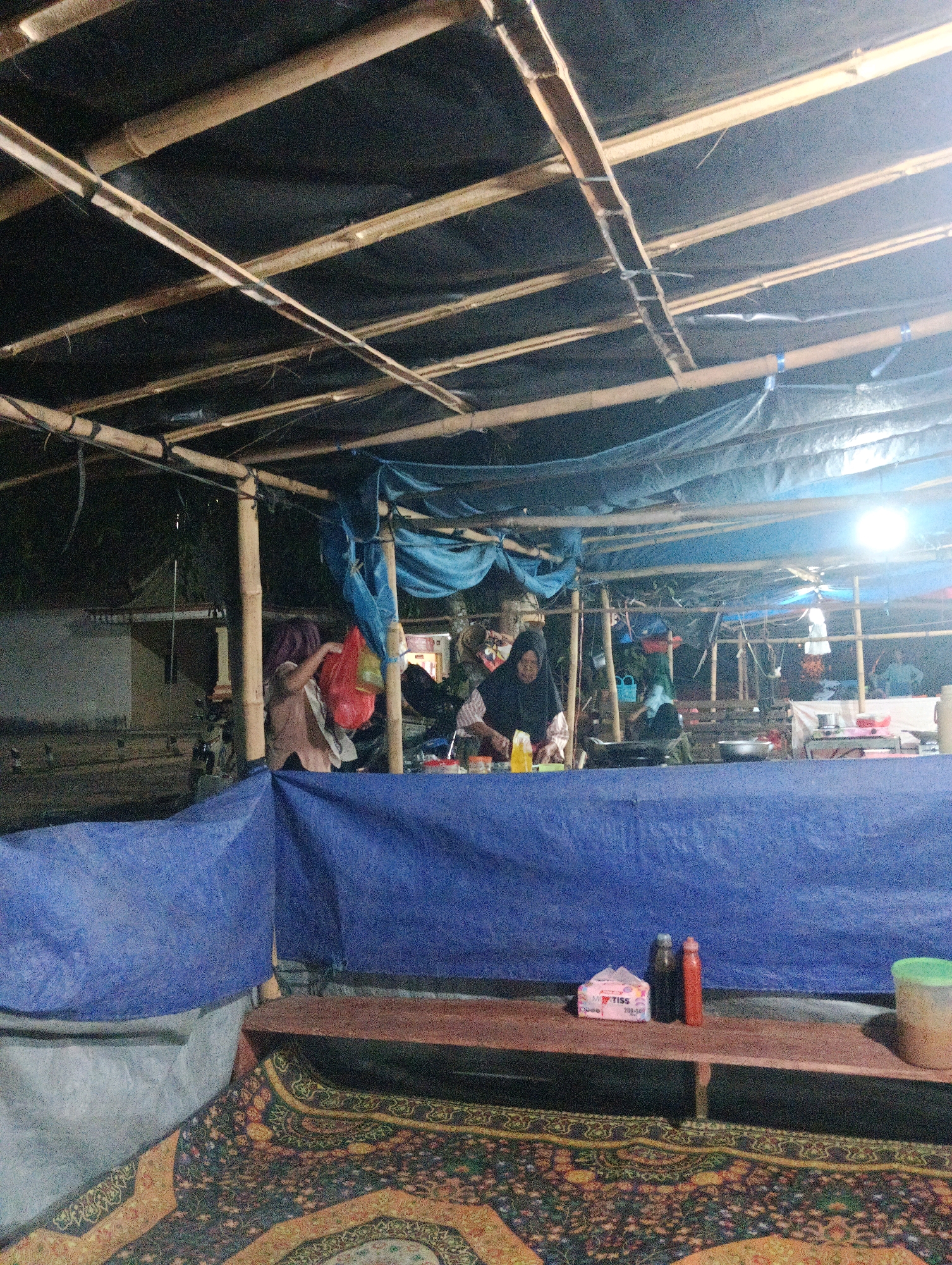 The image depicts an outdoor stall or eatery, possibly in Pematang Jaya, Indonesia. The structure is constructed with bamboo poles and covered with a dark tarp and blue tarpaulin, suggesting it is a temporary or makeshift setup. In the background, there are glimpses of buildings and trees, indicating an urban or semi-urban environment.
In the foreground, a wooden plank acts as a counter, displaying a packet of tissues labeled "TISS," two bottles (one dark, one orange), and a light green container. A patterned rug covers the ground in front of the counter.
The middle ground shows two women behind the counter. One woman, wearing a dark hijab and a patterned shirt, is actively preparing food, with various containers and utensils visible on the counter. Another woman, wearing a light brown shirt and a headscarf, is handling a red plastic bag, possibly containing groceries. There are more cooking supplies and equipment in this area. A motorcycle is parked to the left of the stall.
The overall scene suggests a lively marketplace or food stall operation, likely during the evening or at night, given the artificial lighting from a bright lamp illuminating the area.