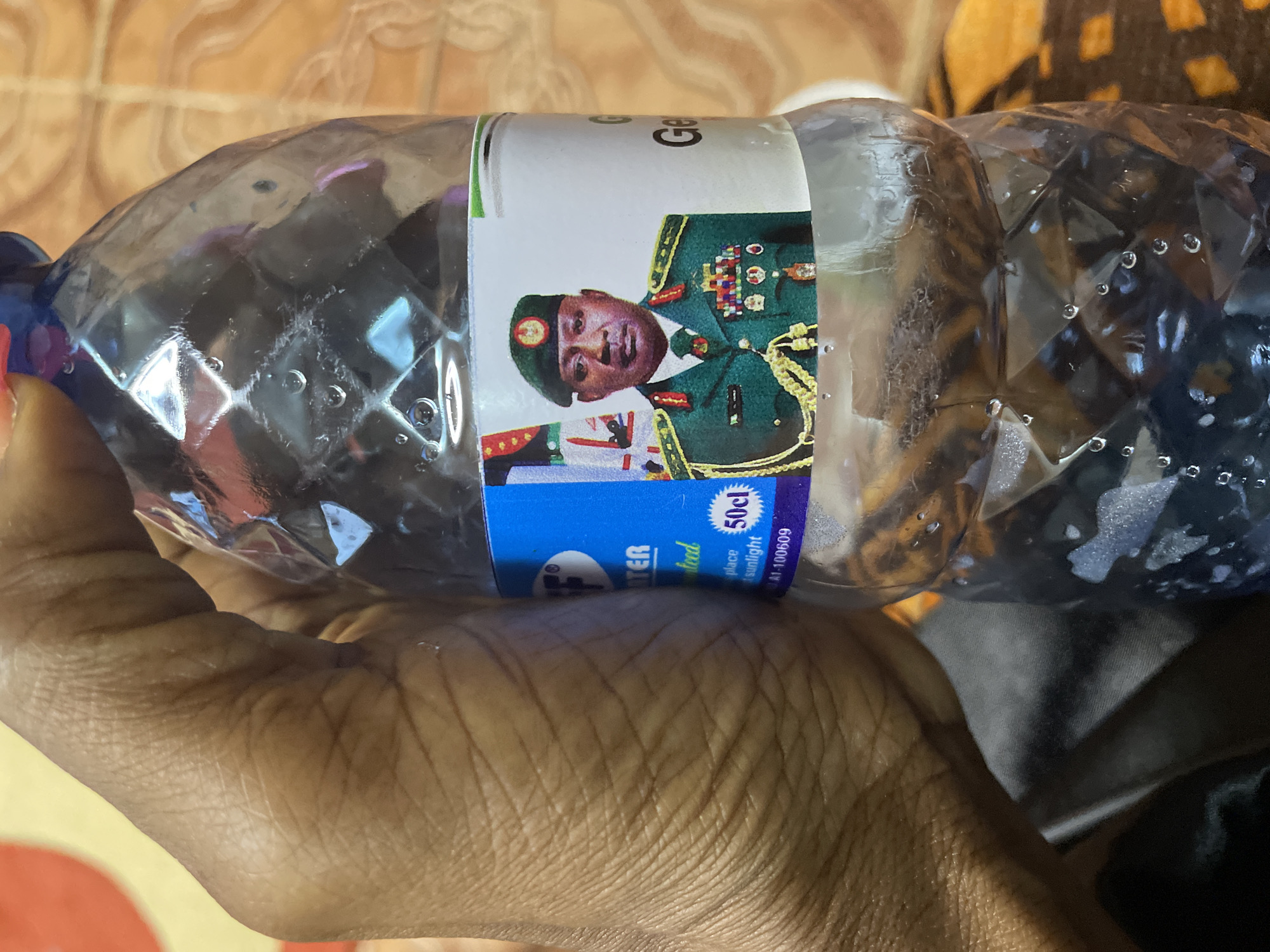 A close-up shot shows a hand holding a clear plastic water bottle. The bottle has a label with an image of a man in a military uniform, wearing a green beret and medals, alongside a flag. Text on the label includes "WATER," "50cl," and what appears to be a serial number. The background shows a patterned surface, possibly a table or mat, in warm tones. The image is taken in Narayi, Nigeria.