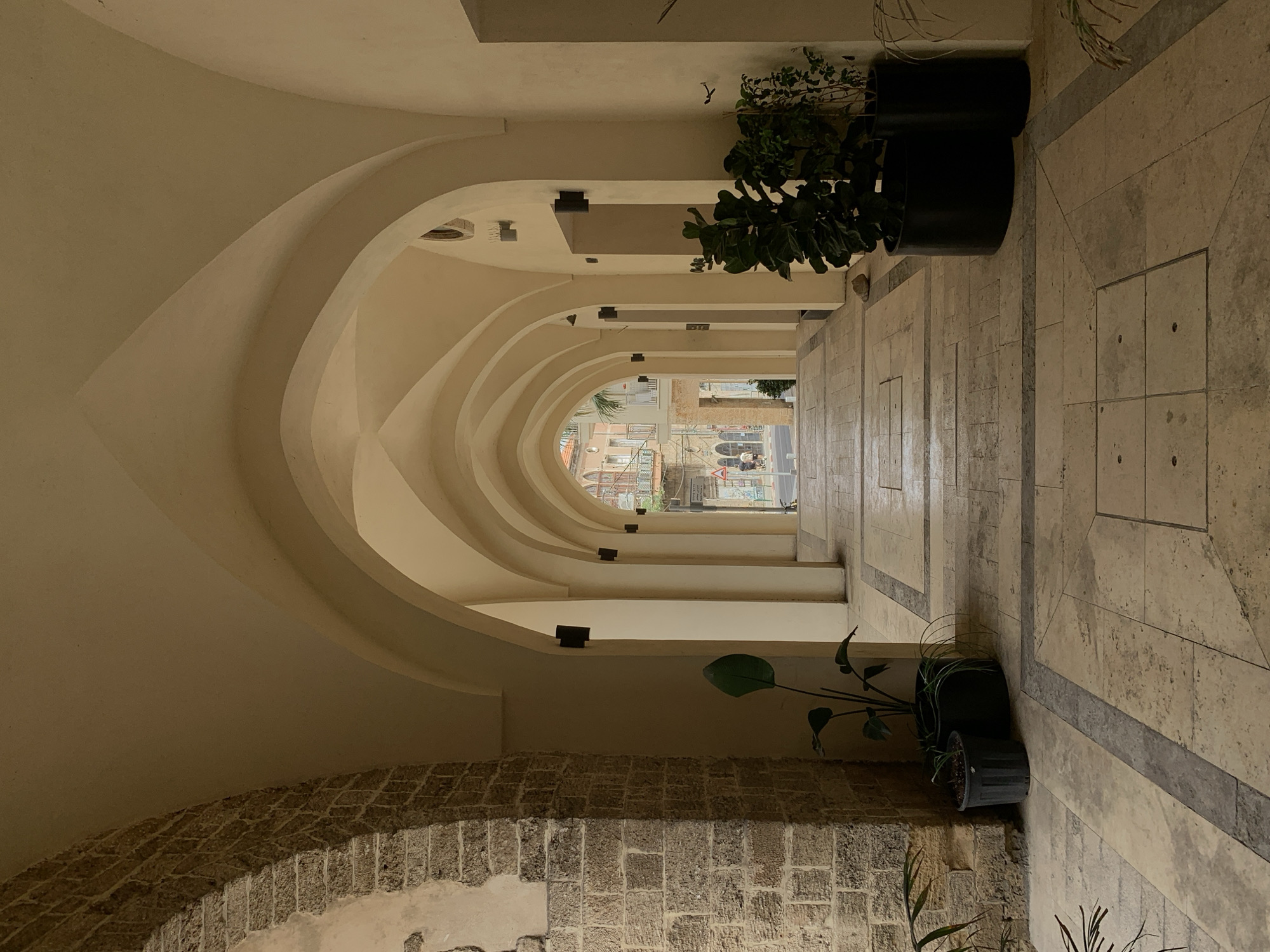 The image depicts a covered walkway with a series of arches, likely in Jaffa, which is part of the city of Tel-Aviv, Israel. The arches, constructed from light-colored stone or plaster, recede into the distance, creating a tunnel-like effect. At the end of the walkway, a street scene is visible, including buildings, traffic, and pedestrians. The walkway itself is lined with plants in dark pots on both sides. The floor is made of light-colored tiles with dark gray inlays. The lower left of the frame includes a wall made of stacked stone bricks.