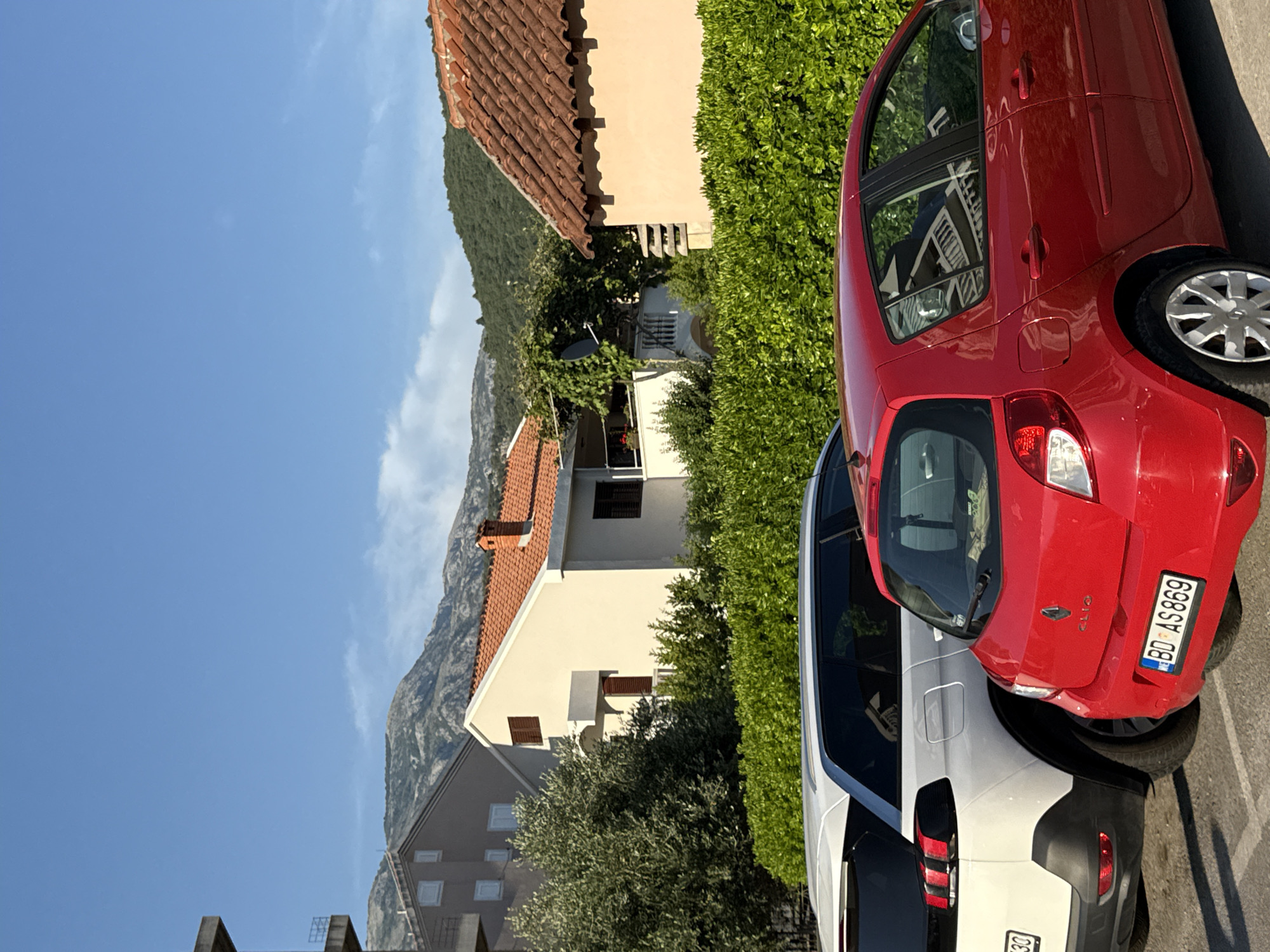 A red Renault Clio and a white SUV are parked side-by-side on a street in Budva, Montenegro. Behind the cars is a dense green hedge, and beyond that, several houses with terracotta tile roofs are visible. In the background, a mountain range rises against a clear blue sky. The scene is captured in daylight, suggesting it is morning or afternoon. The cars are the primary focus, with the red Clio positioned in the foreground and the white SUV partially visible to its left. The license plate of the red car reads "BD AS 869". The overall setting appears to be a residential area in Budva, likely a coastal town, given the presence of mountains and vegetation.