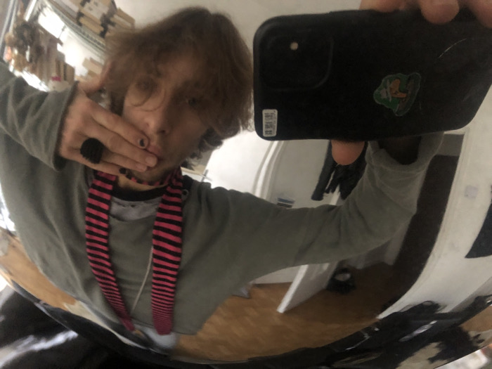 The image captures a selfie taken by an individual in a highly reflective, distorted mirror, reflecting an indoor setting in Paris, France. The subject has shoulder-length brown hair and is wearing a light grey long-sleeved shirt over a darker inner garment, paired with a black and pink horizontally striped neck accessory. The right hand, adorned with a dark, textured ring on the index finger and black-painted fingernails, covers the lower portion of the subject's face. A black smartphone, featuring a green and brown sticker and a white label, is held by the subject's left hand to capture the image. The distorted background reveals shelving units stocked with items, possibly boxes or merchandise, in the upper left quadrant, and a white architectural element, likely a doorway or wall, on the right. A light-colored floor is visible at the bottom of the reflection.