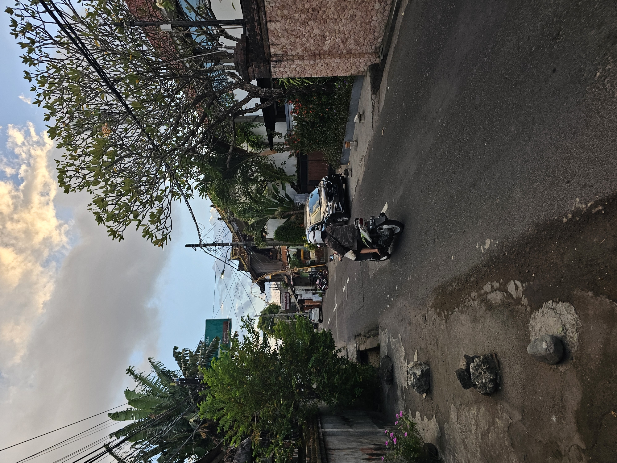 A motorcyclist wearing a camouflage jacket and a white helmet rides down a paved street in Denpasar, Indonesia. The street is lined with trees and buildings, some of which are multi-story. A black car is parked on the right side of the road. A green sign with Indonesian text is visible on the left side of the street. The sky is cloudy with patches of blue. The scene appears to be a typical urban street in Indonesia, likely a residential or commercial area.