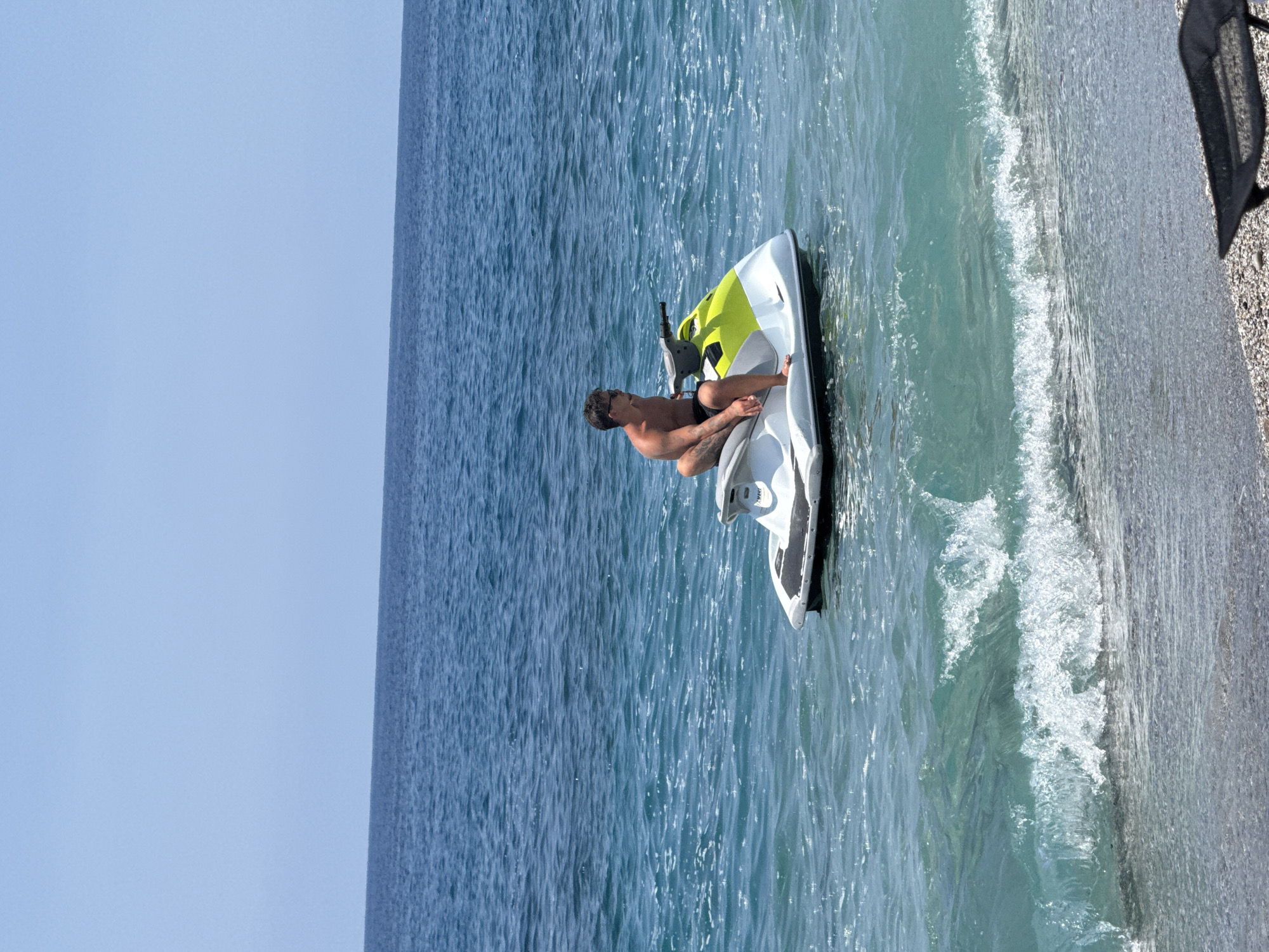 A sunlit coastal scene in Alanya, Turkey, depicts a man on a personal watercraft near a pebbled beach. The shirtless man, with dark hair and wearing sunglasses, is seated on a white and lime green jet ski. He wears dark swim trunks and exhibits visible tattoos on his right arm. He is looking towards the right side of the frame. The personal watercraft is stationary, floating in the clear, light blue-green water close to the shore. The sea gradually transitions from this lighter hue near the beach, where small waves break on the coarse pebbled surface, to a deeper blue further out towards the horizon. The sky above is clear and uniformly light blue, indicating bright, sunny weather. A small, black, foldable beach chair or similar object is partially visible on the beach in the lower right corner of the frame.