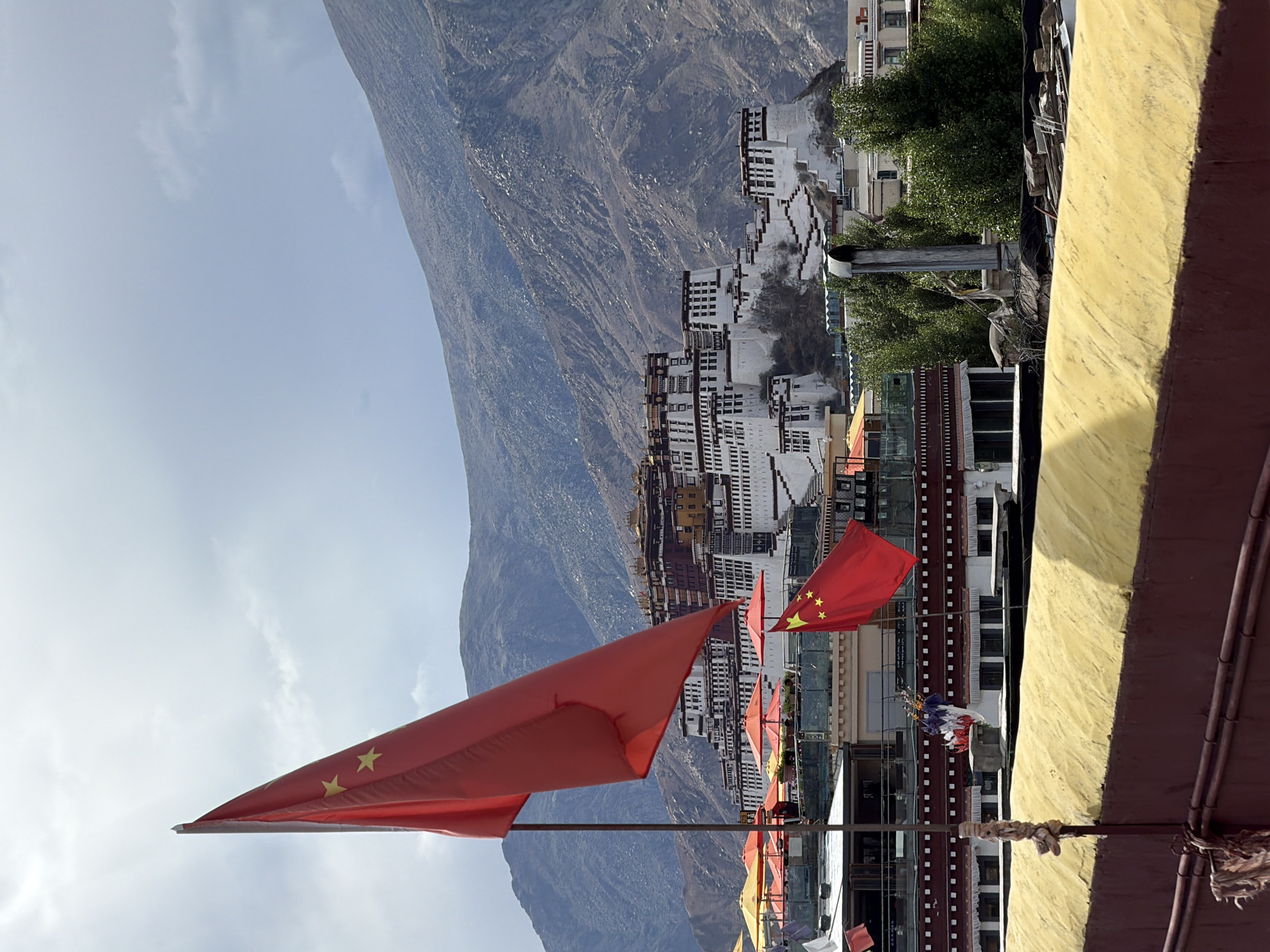 This image captures a vibrant and grand landscape, showcasing a settlement nestled at the base of imposing mountains under a bright sky. In the foreground, a large, prominent red flag, identified as the national flag of China by its five yellow stars, unfurls diagonally towards the upper left. Several other smaller red flags and some multi-colored prayer flags (blue, white, red, green, yellow) are also visible in the mid-ground, attached to poles or lines, suggesting a festive or sacred atmosphere.
To the right, a textured yellow wall and dark brown structural elements, possibly part of a building's exterior or rooftop, form the immediate foreground. Beyond this, a series of multi-story buildings, predominantly white with dark windows and some featuring traditional brown accents, stretch across the mid-ground. These structures exhibit an architectural style that appears historical and culturally specific, possibly hinting at a monastery or traditional settlement. Green trees are interspersed among the buildings, adding touches of nature to the urban environment.
Dominating the background are majestic, rugged mountains, their slopes covered in rock formations and sparse vegetation, suggesting a high-altitude or arid region. The sky above is a clear light blue with a few wispy white clouds, indicating fair weather and bright daylight, likely late morning or early afternoon.
Given the presence of the Chinese flag, the traditional architecture, and the mountainous setting, particularly with the context of "Chengguan District, China," the location is strongly suggested to be in Tibet, possibly Lhasa, known for its unique blend of traditional Tibetan buildings and Chinese presence amidst the Himalayas. There is no discernible activity or event taking place, and no visible text apart from the flag's design. The image evokes a sense of cultural heritage and natural grandeur.