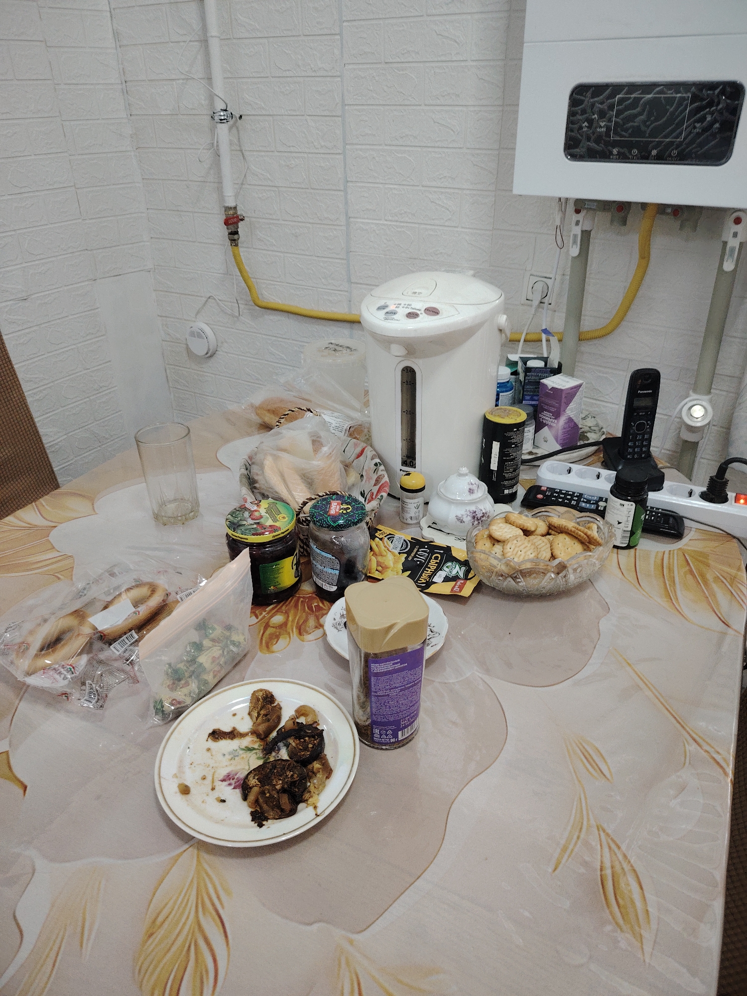 The image depicts a table laden with various food items and household appliances, possibly in a kitchen or dining area. On the table, there are jars of jam, a bowl of cookies, bread products, a plate with remnants of a meal, and a container of instant coffee. A white electric kettle is prominent, along with a cordless phone and several power strips and cords. The background reveals a white brick-patterned wall with visible plumbing and a gas line, suggesting a utility area. A white gas water heater is mounted on the wall. The overall scene suggests a casual meal or gathering. The city of Astana, Kazakhstan, is not directly referenced or visually identifiable in the image.