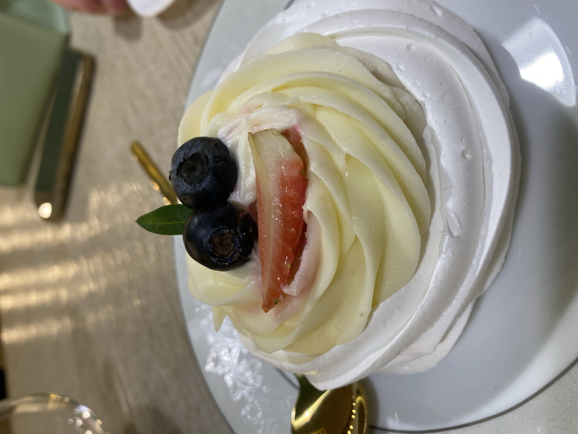 The image displays a close-up of a single dessert served on a white, round ceramic plate in Izhevsk, Russia. The dessert features a base of white, textured meringue, piped into a circular shape. On top, pale yellow cream is piped in concentric swirls. The dessert is garnished with two dark blue blueberries placed at the upper center, a horizontal slice of red strawberry positioned beneath them, and a single green leaf situated above and between the blueberries. A golden spoon is visible on the plate to the left of the dessert. The background, out of focus, reveals a light-colored table surface. Partially visible objects include a clear glass on the upper left, a smartphone with a light green case on the upper right, and a human hand next to a small white dish on the far right. The environment appears to be an indoor setting, consistent with a cafe or restaurant.