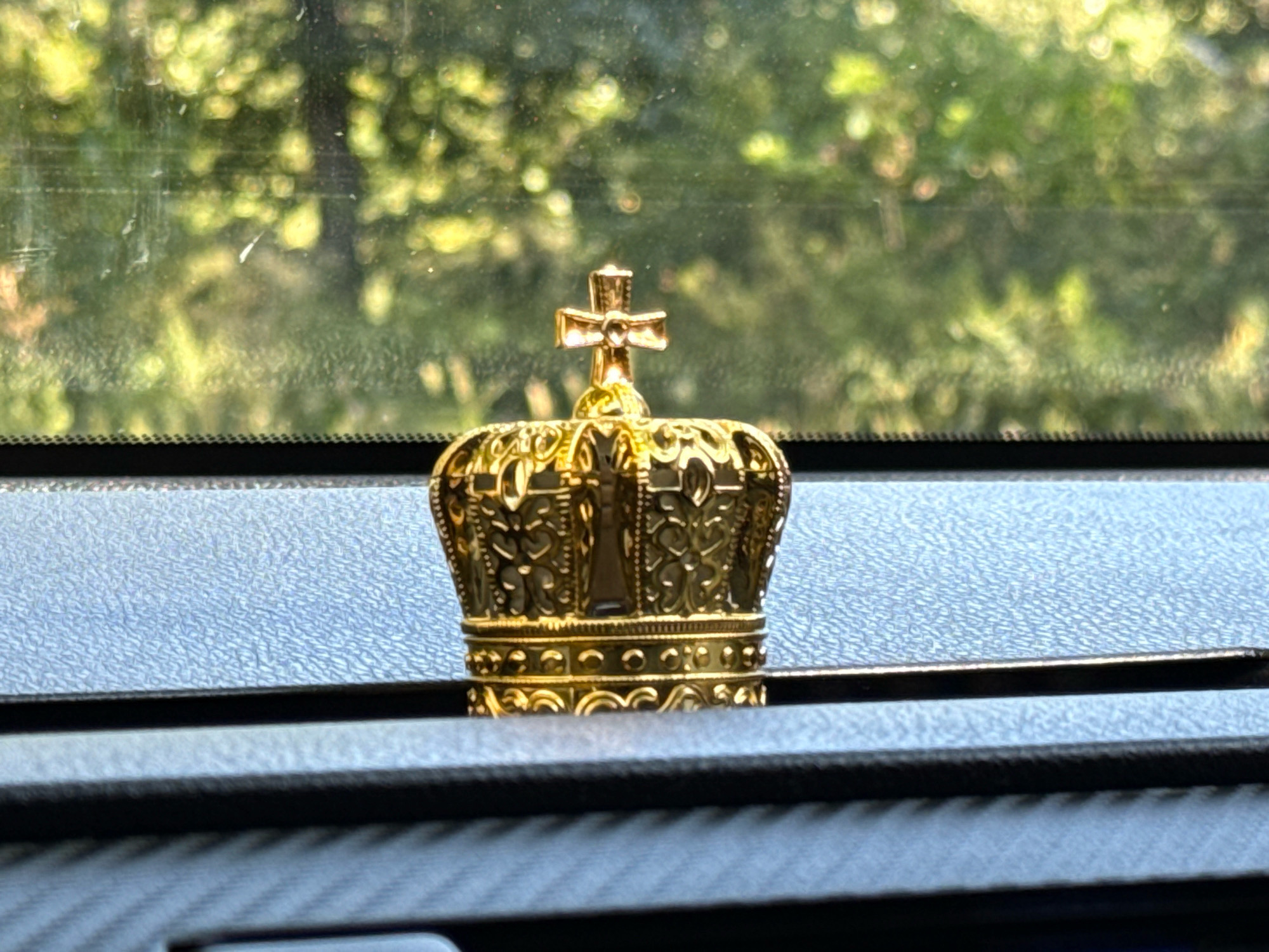 A close-up, eye-level, medium shot captures a golden crown-shaped ornament resting on a textured, dark gray dashboard. The ornament, featuring intricate filigree and a cross on top, is centrally located and in sharp focus. Behind the ornament, a car windshield is visible, showing a blurred backdrop of lush green foliage, illuminated by sunlight. The bokeh effect of the background suggests it is out of focus. The scene appears to be indoors, likely within a vehicle, with the dashboard and windshield indicating the car's interior. The lighting is natural, casting a warm glow on the golden ornament. There are no people present in the image.
