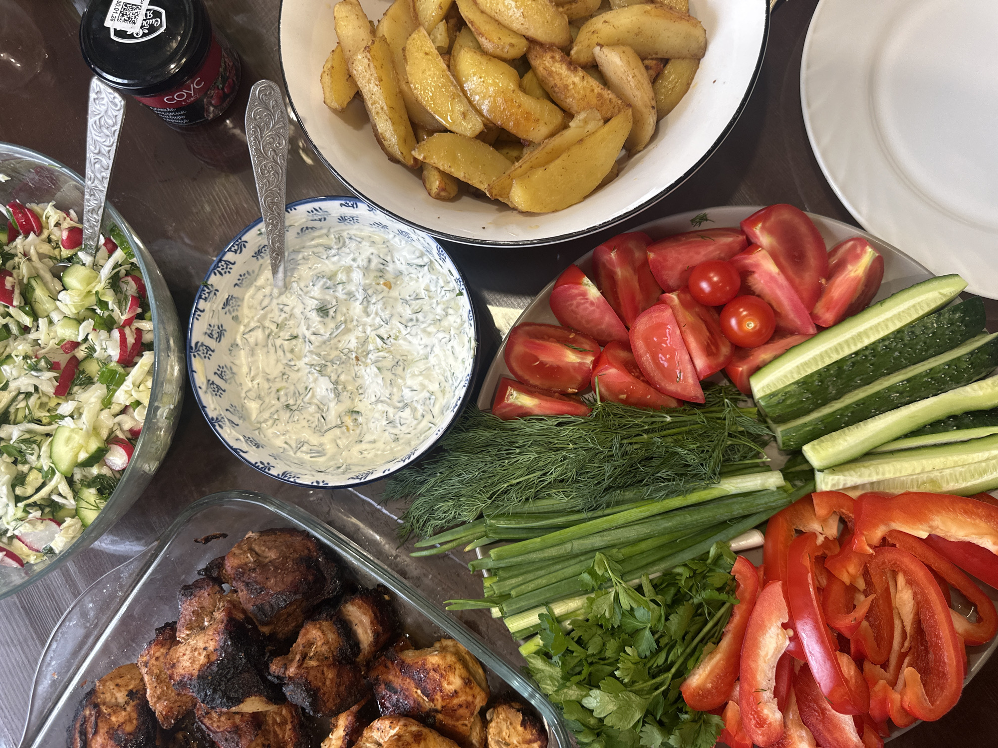 A top-down view of a table laden with food, presumably taken in or near Chaikovsky, Russia, given the Cyrillic script on the jar. Dishes present include: a bowl of fried potato wedges, a bowl of salad with radish and cucumber, a bowl of dill sauce, a platter of sliced tomatoes, cucumbers, red peppers and herbs and a platter of grilled chicken. The table surface appears to be dark brown. A jar of sauce and white plates are visible near the top of the frame. Silver serving spoons are present in the salad and the dill sauce.