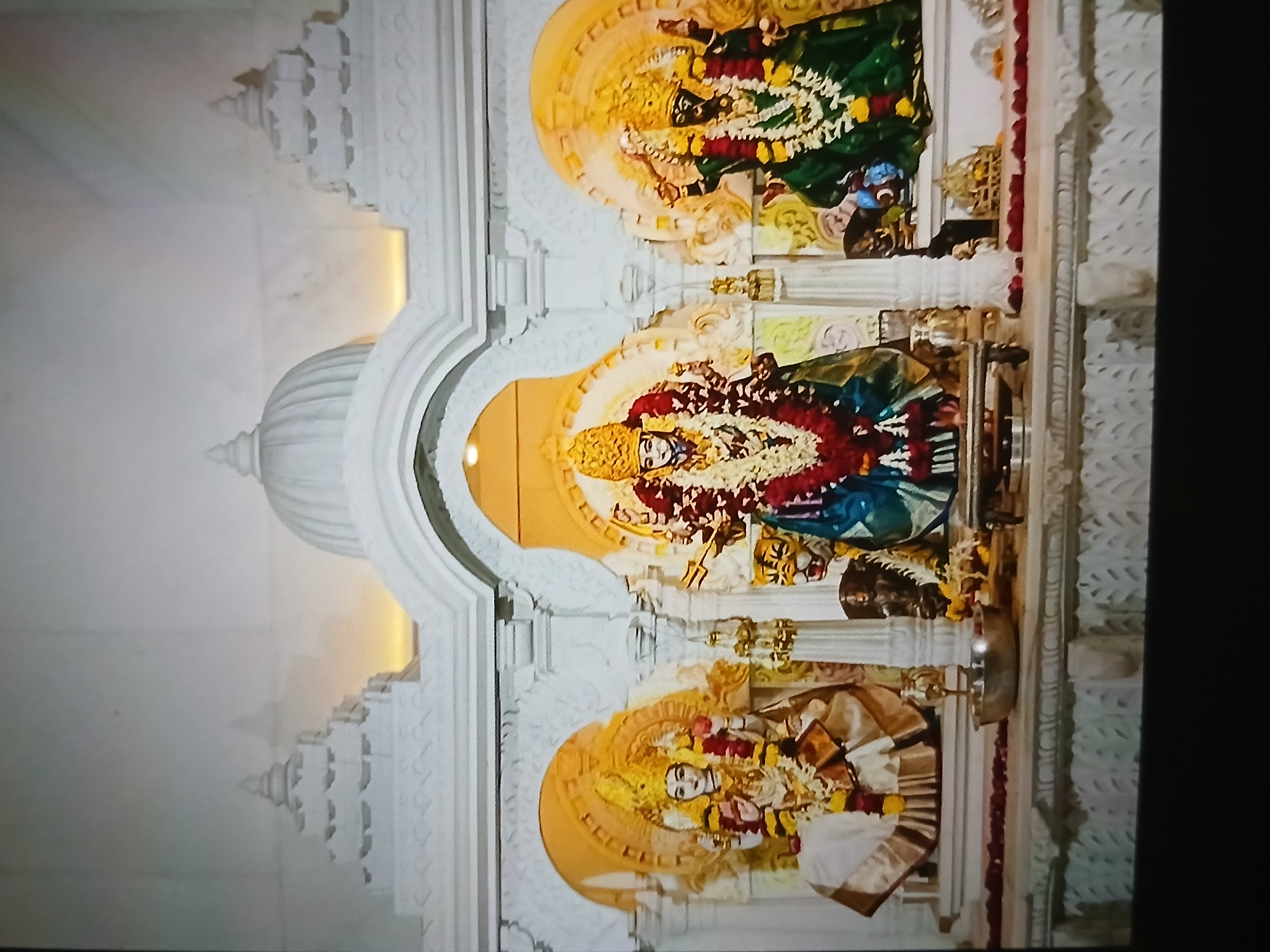 The image depicts a Hindu shrine, likely located in Bengaluru, India, given the architectural style and religious iconography. The shrine features three deities displayed in individual niches, elaborately decorated with garlands and adorned with gold ornaments. The architectural structure is white, adorned with intricate carvings and detailing, and crowned with a dome-shaped structure at the top. The color palette is dominated by gold, white, and vibrant colors of the deity's attire and floral offerings. The photo is taken in low light, with artificial lighting highlighting the deities and the architectural details.