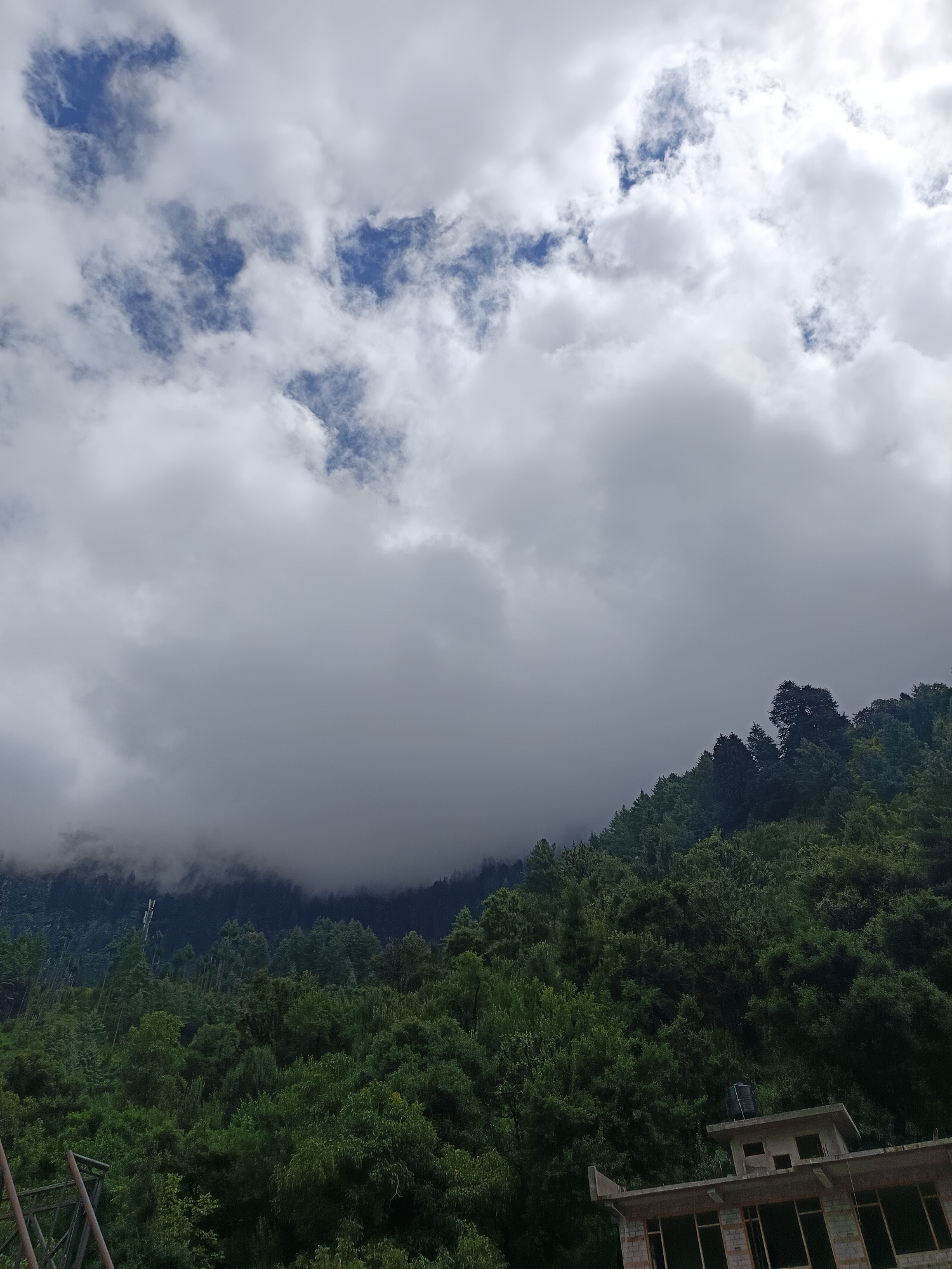 The image captures a mountainous landscape with a dense forest covering the slopes. A building, partially constructed with a water tank on its roof, is visible in the lower right corner, suggesting it is located on the outskirts of a populated area. The sky is overcast with thick clouds, partially obscuring the higher mountain peaks. The presence of lush green trees indicates a humid climate, typical of regions like Vashisht, India, known for its natural beauty and spiritual significance. No people or animals are present in the frame. The scene is static, with no apparent activity.