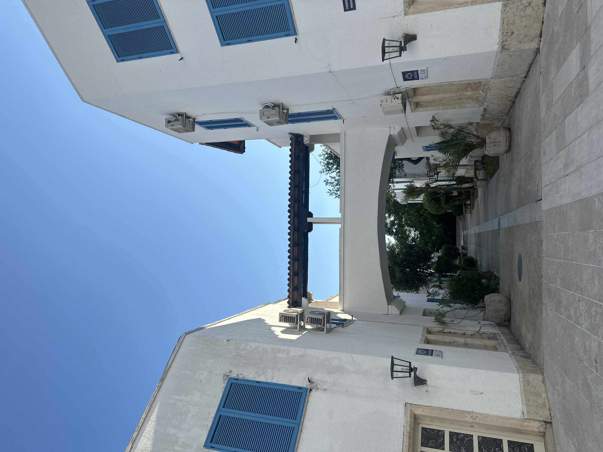 The image depicts a bright, sunny day in what appears to be a Mediterranean coastal town, likely Budva, Montenegro, given the location context. The scene is dominated by traditional whitewashed buildings with blue shutters on the windows. The perspective is unusual, with the buildings seemingly tilting upwards, and the sky occupying a significant portion of the frame.
In the foreground, a white building with blue shutters is visible, with an air conditioning unit mounted on its exterior. Further back, an archway leads to a walkway lined with more white buildings and potted plants. Another air conditioning unit is mounted on a building near the archway. On the wall of one building, a black lantern is affixed, and a sign with blue lettering is visible. Another sign, also with blue text, reads "POLA LOKVA" and has an arrow pointing left. The ground is paved with light-colored stone tiles.
In the background, through the archway, dense green foliage and trees are visible, suggesting a well-maintained outdoor area or courtyard. The clear blue sky indicates fair weather, likely midday or early afternoon due to the bright lighting and shadows. There are no people visible in the image. The overall impression is one of a tranquil, picturesque setting.
