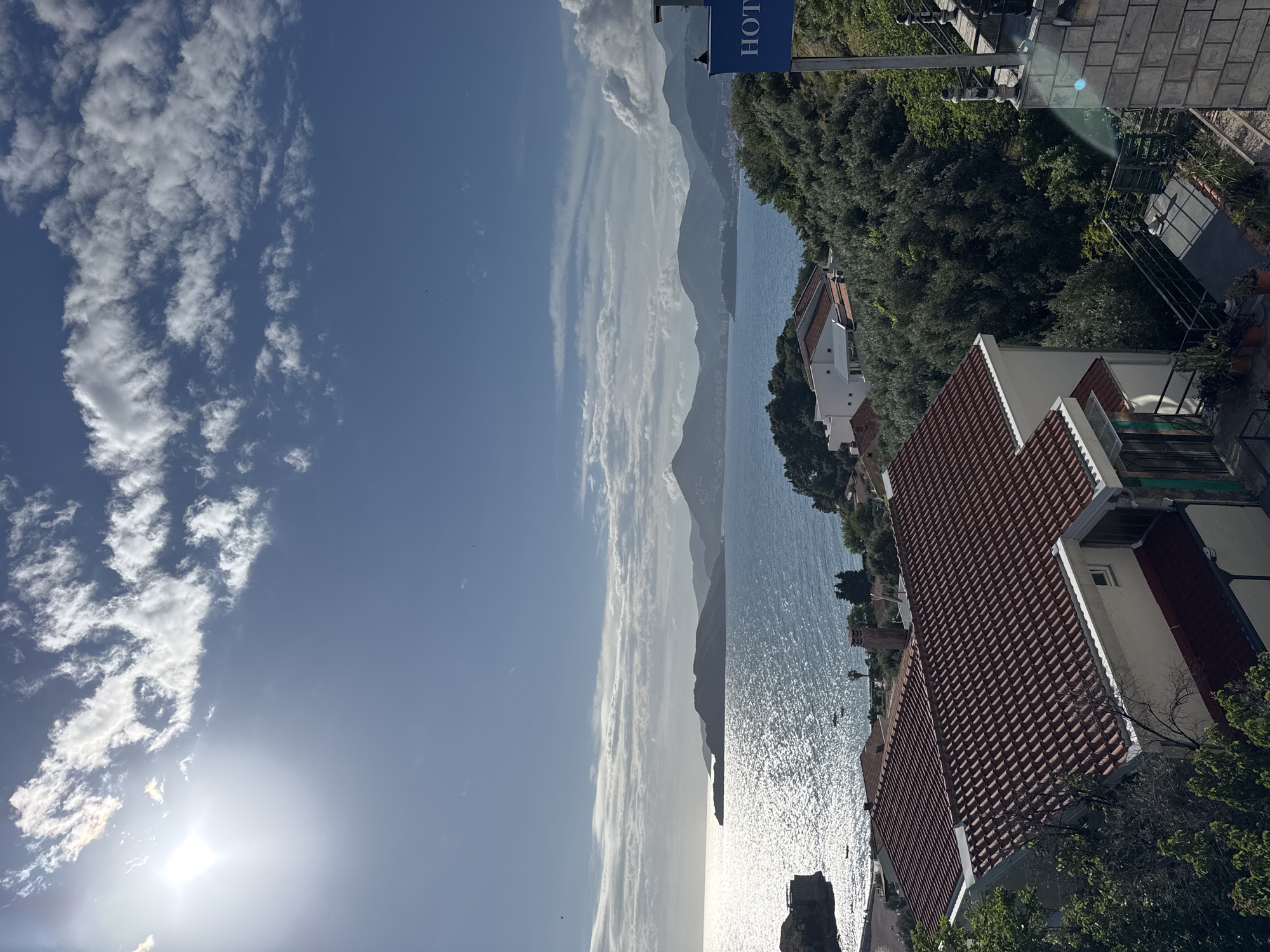 Captured from an elevated vantage point in Sveti Stefan, Montenegro, the image presents a panoramic view. The foreground features buildings with terracotta tiled roofs partially obscured by lush greenery. Beyond lies the Adriatic Sea, its surface reflecting the bright sunlight creating a shimmering effect. In the distance, imposing mountains form a hazy backdrop. The sky, a gradient of blue, is adorned with scattered clouds, and a "HOTEL" signpost peeks into the frame on the right.
