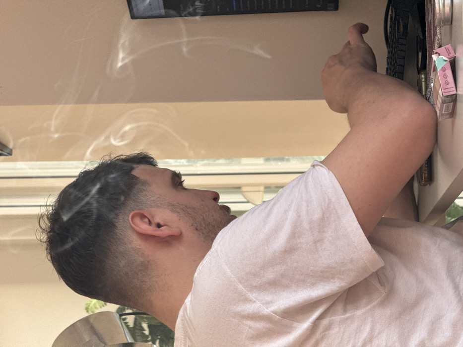 An indoor scene within Vozdovac Urban Municipality, Serbia, depicts a male individual seated at a light-colored desk, viewed from his left-side profile. The male, appearing to be a young adult, has short, dark hair with a faded haircut and stubble. He wears a light-colored short-sleeved t-shirt. His right arm is bent, and his hand rests on a black computer keyboard, indicating interaction with a computer. A partially visible black computer monitor displaying text on a dark background is positioned to his right. On the desk, a metallic-cased smartphone, an open light pink cigarette pack, and a small, silver-colored metal tin are visible. White smoke is clearly observed circulating in the air above and around the individual's head, suggesting active smoking. Behind the individual, a window or glass door allows natural light into the room, revealing a glimpse of green foliage outside. The background wall is a solid, light beige color.