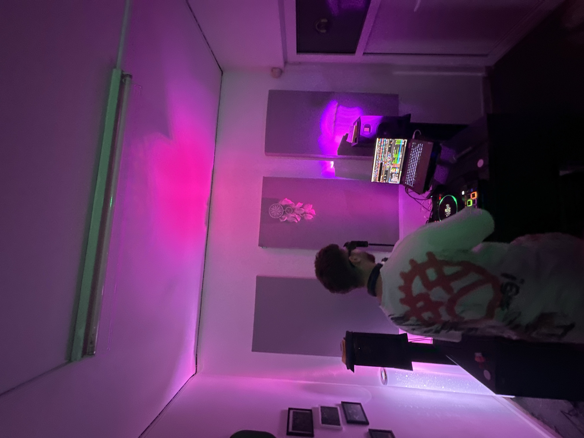 The image shows an indoor music studio or DJ booth located in Plovdiv, Bulgaria. A male subject is visible from the back, positioned at a DJ setup. He wears a white t-shirt with a large red graphic design on the back and dark-rimmed glasses, with headphones resting around his neck. The DJ equipment comprises a black console with illuminated green and multi-colored controls, and an open laptop displaying music production software, situated on a stand to the right of the console. Two black studio monitor speakers flank the DJ table. The room's white walls are fitted with multiple rectangular gray soundproofing panels. A white dreamcatcher with pink tassels is affixed to a central panel on the back wall. Framed pictures are arranged vertically on the left wall. A tall, cylindrical lamp with internal sparkling elements stands to the left of the DJ setup. Overhead, a single fluorescent light fixture emits a green hue. The primary illumination in the room is provided by ambient magenta and purple lighting, casting prominent colored reflections. The floor is dark, and a white door frame is visible on the far right.