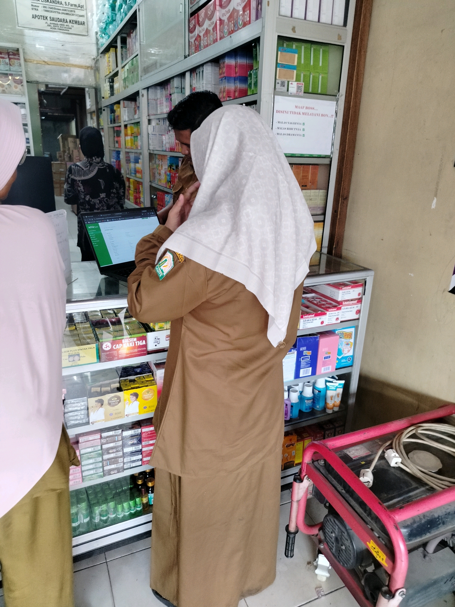 In the city of Perapattitipanjang, Indonesia, an indoor scene depicts the interior of a pharmacy. Three individuals are present. One person, wearing a pink headscarf and brown pants, stands in the foreground on the left, partially obscuring a laptop. To the right of this person, another individual is seen from behind, wearing a brown uniform with an emblem on the sleeve and a light-colored headscarf. They are engaged with the laptop. A third individual, wearing a black headscarf and patterned clothing, is visible in the background, to the left of the laptop. Shelves stocked with various medicinal products and personal care items line the walls and are displayed in glass cabinets. A red generator is situated in the lower right portion of the frame. The overall environment suggests a commercial establishment, likely a pharmacy or drugstore.