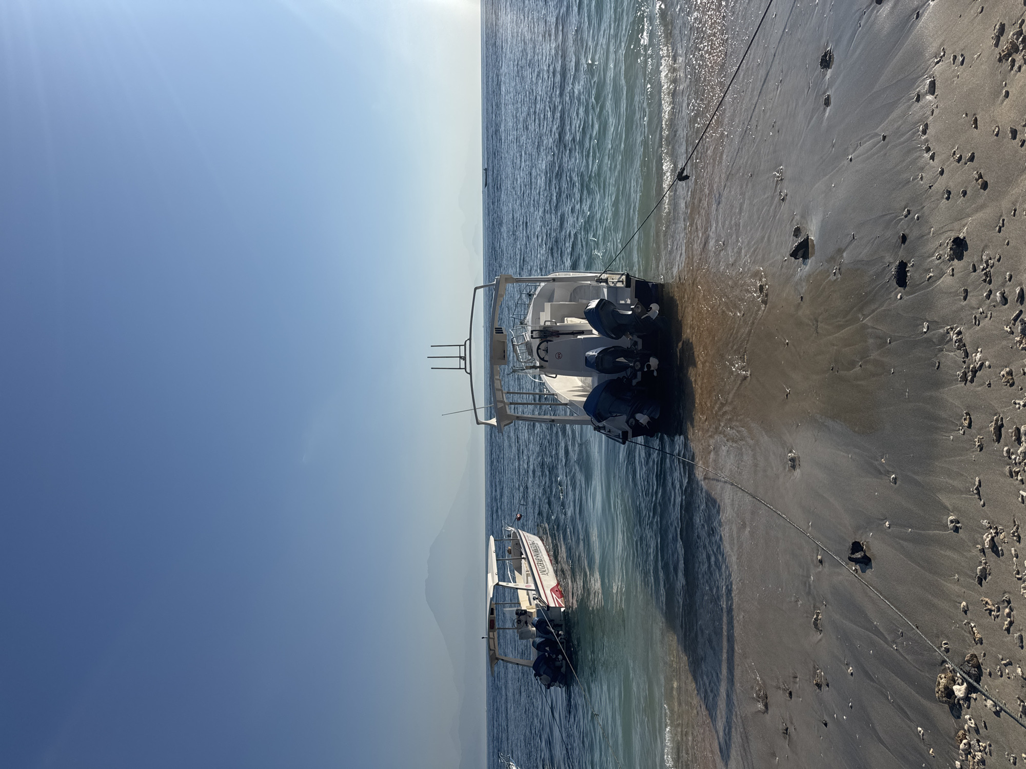 The image depicts a sunlit coastal scene in Nusa Penida, Indonesia. In the foreground, dark, wet sand exhibits ripples from receding waves and is scattered with numerous small pieces of light-colored coral debris and dark rocks. A thick black mooring rope runs diagonally from the lower center of the frame, disappearing into the water to secure the nearest boat.
Two powerboats are visible near the shore. The boat on the right, partially beached on the wet sand and partly floating in the shallow water, is white with a canopy structure. It faces away from the viewer, displaying three dark blue outboard engines mounted on its stern. To its left, a second powerboat, white with red trim, floats entirely in the water. The text "ANUGERAH SNORKELING" is clearly visible on its side. This boat also features multiple outboard engines. Both vessels are tethered to the shore by ropes.
The water transitions from light turquoise near the shore to a deeper blue further out, appearing calm with small waves breaking on the beach. The ocean extends to a clear horizon, where a small, distant boat is faintly visible on the right side. In the background, a series of hazy, distant mountains rise, with a prominent, large conical peak (identifiable as Mount Agung on Bali) situated behind the left boat, a characteristic vista from Nusa Penida. The sky is a clear, bright blue, with bright sunlight originating from the upper right, casting long, distinct shadows on the sandy beach and creating a noticeable lens flare in the upper right portion of the sky. No people are visible within the frame.