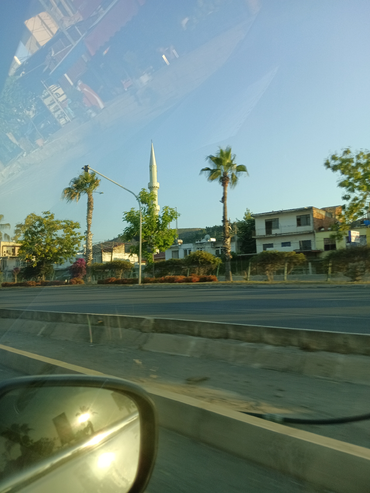 This media file captures a roadside scene in Gazipaşa, Turkey, likely during a car journey. The image is taken from inside a vehicle, as evidenced by the reflection of the side mirror in the foreground, which shows palm trees and buildings.
The main subjects in the scene include a minaret of a mosque, several palm trees, and buildings lining the road. The buildings appear to be a mix of residential and perhaps commercial structures, with some showing signs of age. Greenery, including trees and bushes, is abundant along the roadside, with some flowering plants adding touches of color. A street light pole is visible, indicating an urban or semi-urban setting. The road itself, with its concrete barriers, suggests it is a main thoroughfare.
The time of day appears to be late afternoon or early evening, given the warm, low-angle sunlight that creates bright reflections and casts long shadows. The sky is clear and blue, suggesting fair weather. The overall mood of the image is peaceful and serene, conveying a sense of travel and observation.
No visible text is clearly legible in the image, though there might be some signage on the buildings that is too blurry or distant to decipher. The reflection in the side mirror is a notable detail, showing a reversed image of the surroundings, including the sun's glare. The file appears to be a still photograph.
