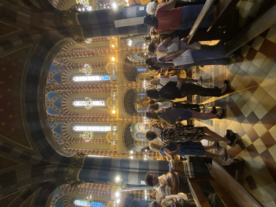 The scene depicts the interior of the Iglesia de Nuestra Señora del Pilar, located in the Recoleta neighborhood of Buenos Aires, Argentina. The environment is a large, ornate church nave characterized by tall, alternating dark and light striped columns and arches supporting vaulted ceilings. The ceilings and upper walls feature intricate patterns in red, gold, and blue, with numerous arched windows containing clear and blue-toned stained glass, each flanked by ornate gold chandeliers. The floor is a checkered pattern of light and dark tiles, reflecting light and casting shadows from the individuals present.
Approximately 20-30 individuals, predominantly adults, occupy the central and right sections of the nave. Most are standing, some observing the interior, while others appear to be waiting or moving slowly. Their gazes are generally directed forward or slightly to the right, towards an unseen focal point. On the far left, a man in a light-colored striped polo shirt is standing, looking down at a mobile phone he holds in his hands. In the foreground, a line of women stands in profile or with their backs partially to the viewer; one woman wears a black and tan patterned dress, and another carries a white tote bag. Wooden pews are visible on both sides of the aisle. The church is brightly illuminated by artificial lighting, creating distinct highlights and shadows.