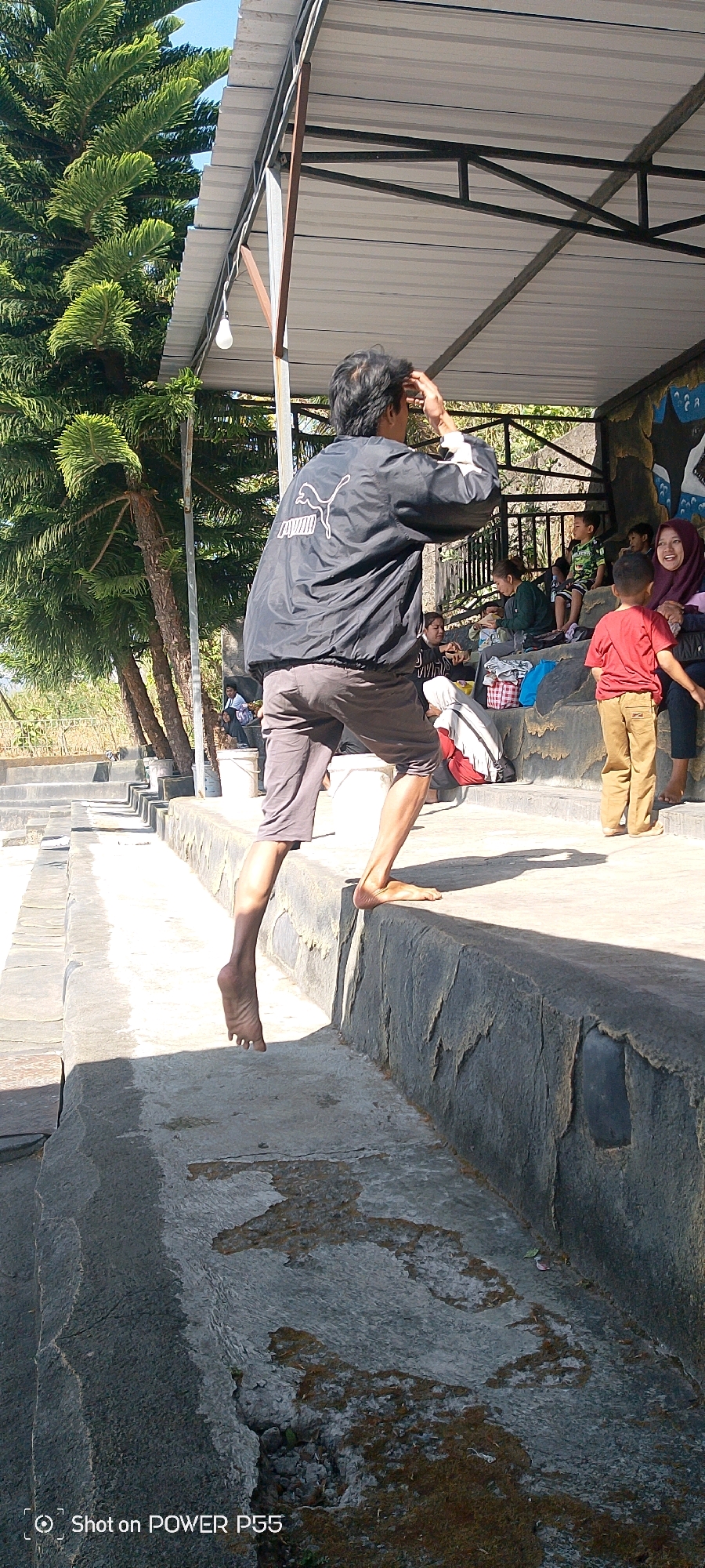 A man in a black Puma jacket and gray shorts is captured mid-jump, his right leg extended as he leaps over a concrete barrier. He appears to be barefoot, with his left foot hovering above the ground. The scene takes place outdoors, with a large evergreen tree on the left and a shaded seating area in the background where several people, including children, are gathered. The ground is made of concrete and appears to be wet in some areas, with a few patches of dirt and debris. The lighting suggests it is daytime, with strong shadows cast by the man and the overhead structure. The background shows a slightly overgrown landscape with a clear sky.