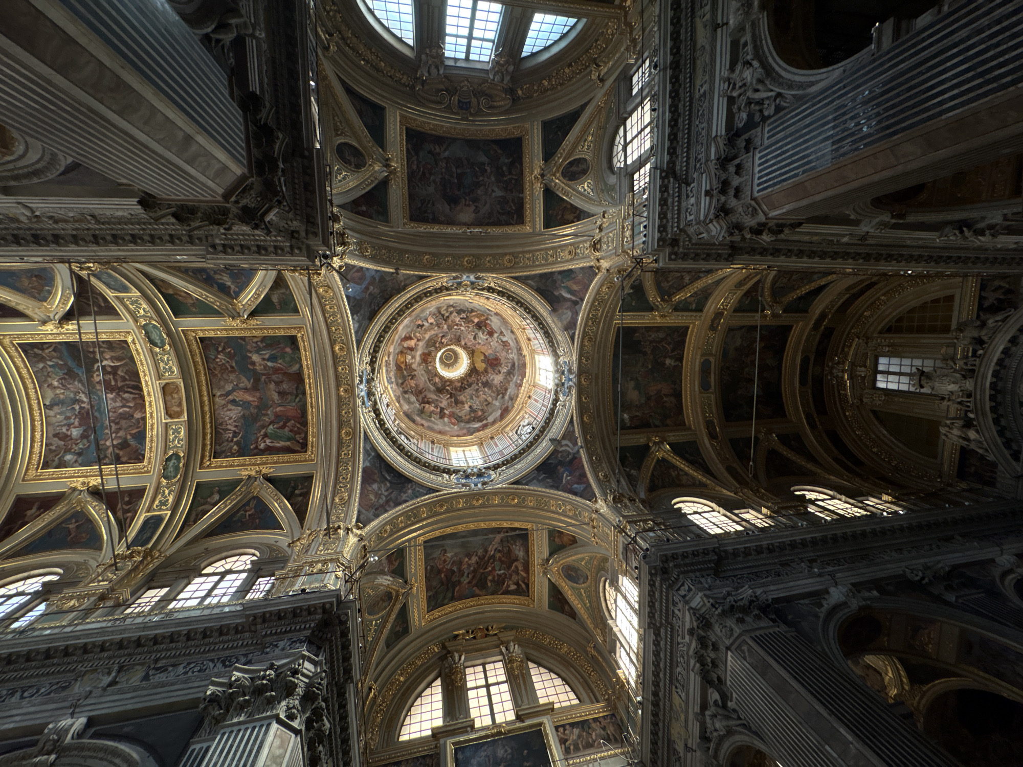The image shows the interior of a church in Genoa, Italy. Looking up, the ornate ceiling features a large dome with paintings, surrounded by smaller panels with more paintings and detailed gold trim. Windows are visible throughout the structure. The architecture is classical, with prominent columns and intricate carvings.