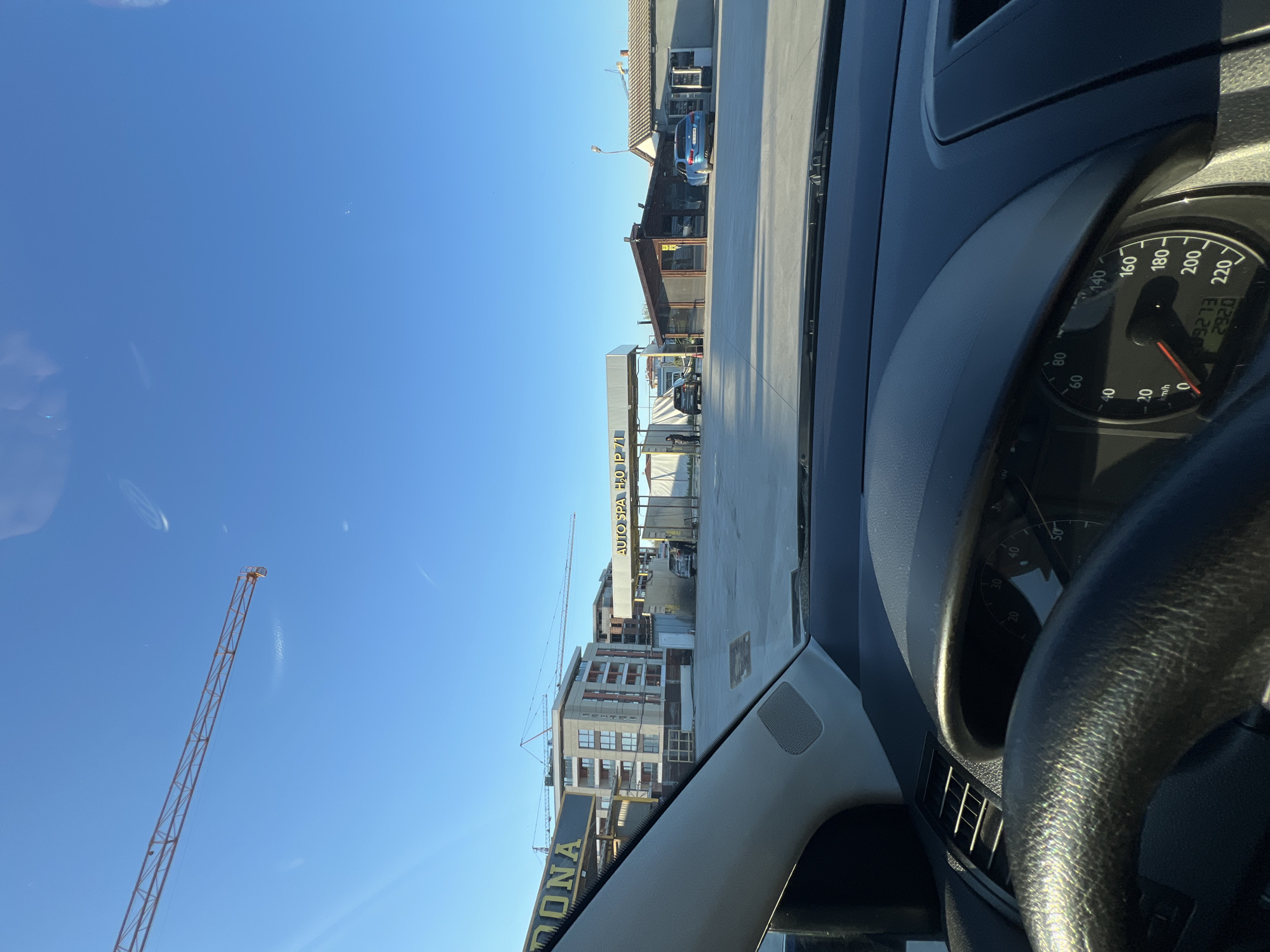 The image, taken from inside a stationary car, shows a bright, sunny daytime scene in an urban or industrial area of Plovdiv, Bulgaria. The car's dashboard is visible in the foreground, with the speedometer at 0 km/h, an odometer reading 3273 km, and a trip meter showing 292.0 km.
Through the windshield, a clear blue sky dominates the left side, featuring a tall, orange construction crane. The mid-ground reveals a paved service area or street lined with various buildings. A prominent sign for "AUTO SPA H₂O IP 71" is visible on one building, suggesting a car wash or service center, with a blue car parked nearby. Another large building, possibly under construction, stands further down, with its multi-story structure and scaffolding visible. A partial sign displaying "DONA" is also seen on the lower left. The overall impression is a vibrant, active area under clear, pleasant weather conditions.