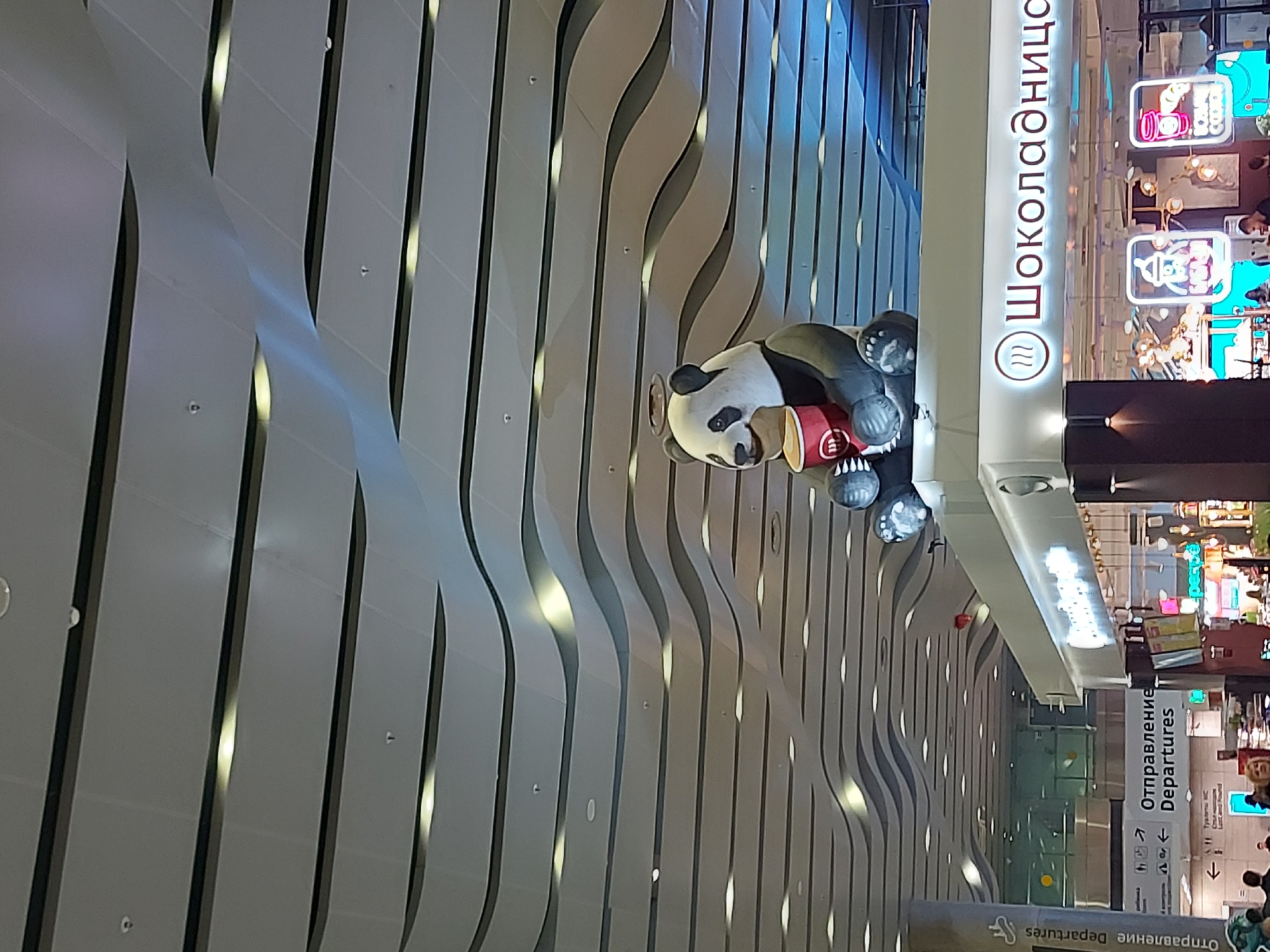 The image captures a scene inside Pulkovo Airport in Saint Petersburg, Russia. A prominent feature is a whimsical display featuring a giant panda holding a coffee cup near a "Shokoladnitsa" cafe. The cafe's signage is visible in Russian Cyrillic script. The airport's interior showcases a modern design with wavy, ribbed wall panels that are evenly spaced and vertically oriented. Signs indicating "Departures" in both Russian and English are also visible, suggesting the image was taken in a terminal area. The foreground features the back of a person's head. Beyond the panda and cafe, there is an open airport space with shops and people.