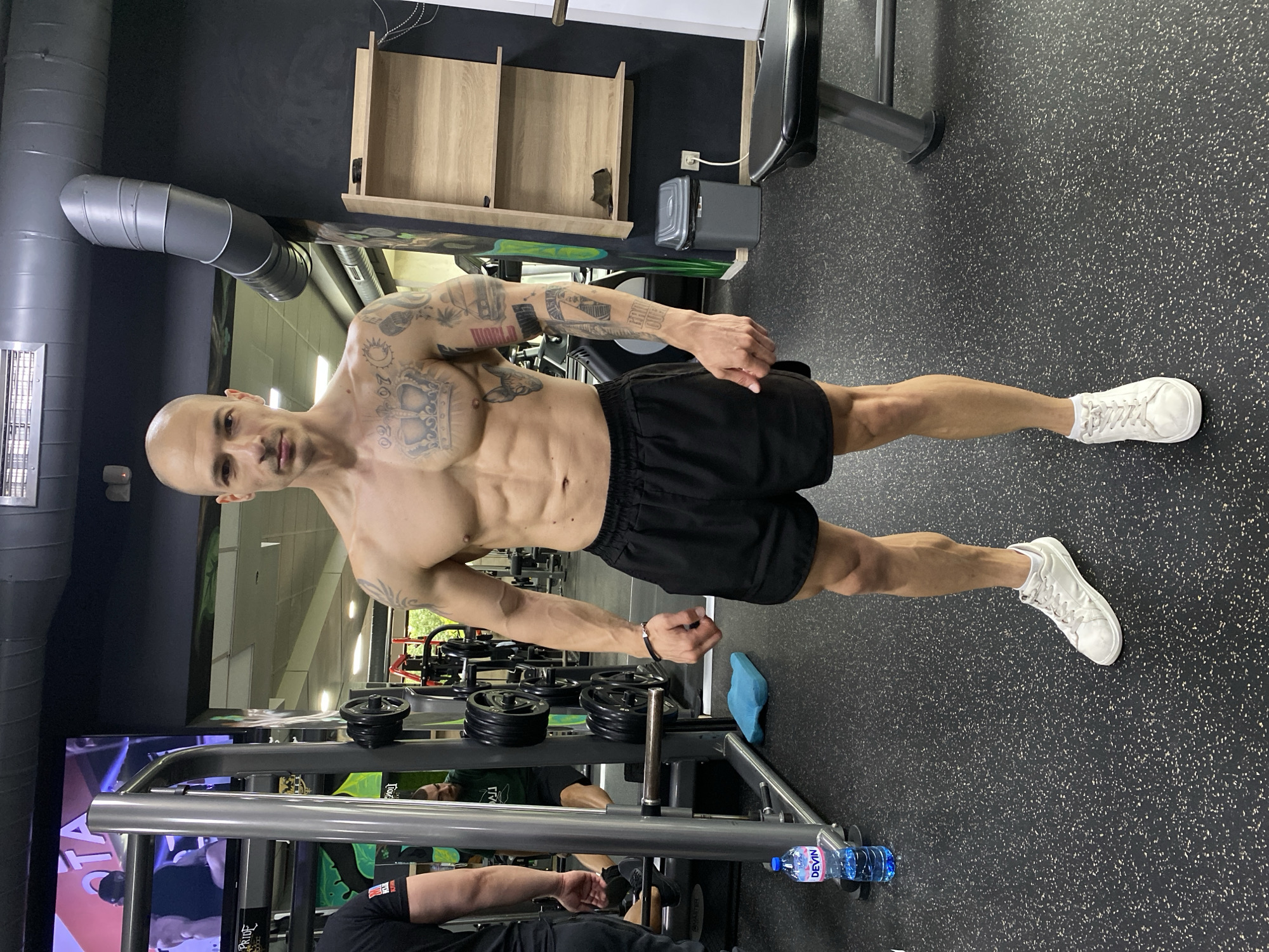 A muscular, bald man with tattoos stands in a gym. He is wearing black shorts and white sneakers, and is shirtless, revealing his toned physique. He is positioned in front of a weight rack loaded with black weight plates. A bottle of "Devin" water lies near the rack. The gym appears to have black rubber flooring with white flecks and a black ceiling with exposed ventilation. The image was captured in the city of Pleven, Bulgaria.