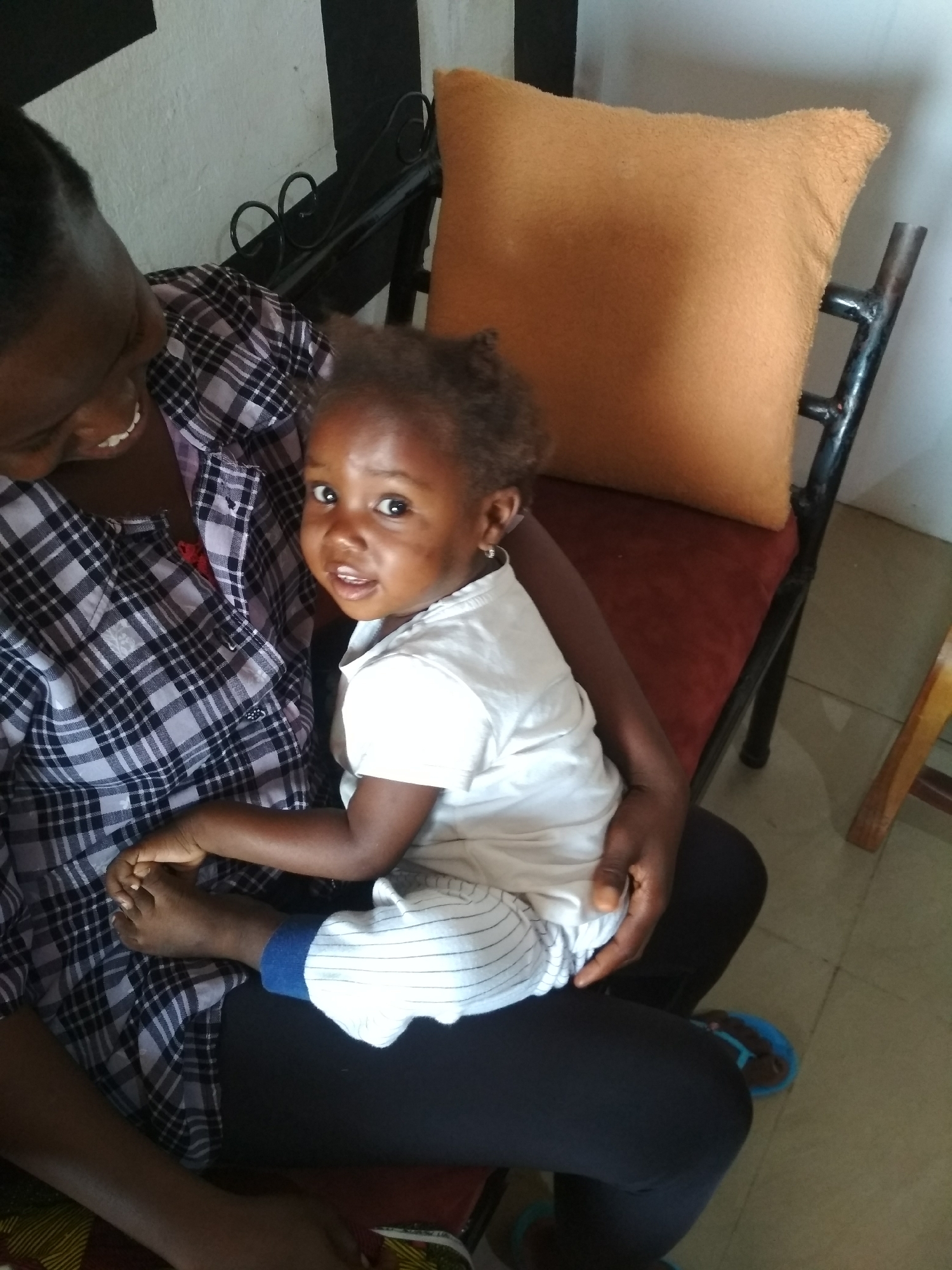 A woman holds a young child on her lap. The woman wears a black and white plaid shirt. The child wears a white shirt and striped pants. The woman smiles at the child. The background includes a brown pillow on a black metal chair, and a black and white striped wall. The location is Unknown, Unknown.