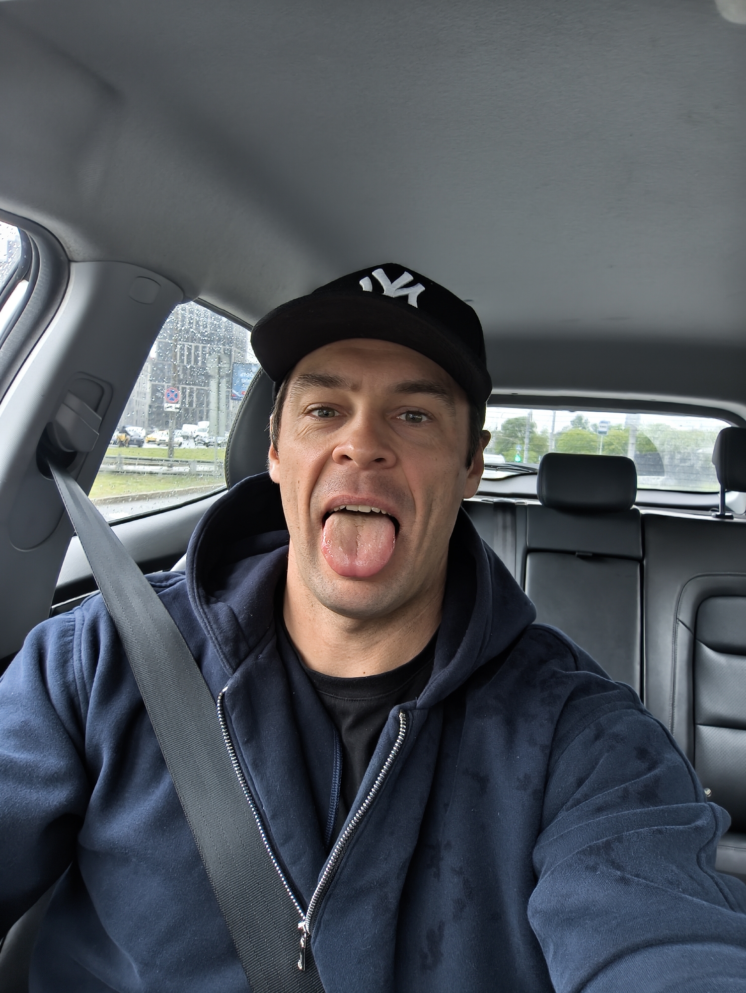 A man is in the driver's seat of a car, wearing a black baseball cap with a white logo, a navy blue zip-up hoodie, and a black t-shirt. He is sticking his tongue out. He is wearing a seatbelt. Rain is visible on the car window. The car's interior has gray and black upholstery. In the background outside, there are buildings and greenery, which, based on the location, are likely Moscow, Russia. A traffic sign is visible through the rain on the window.