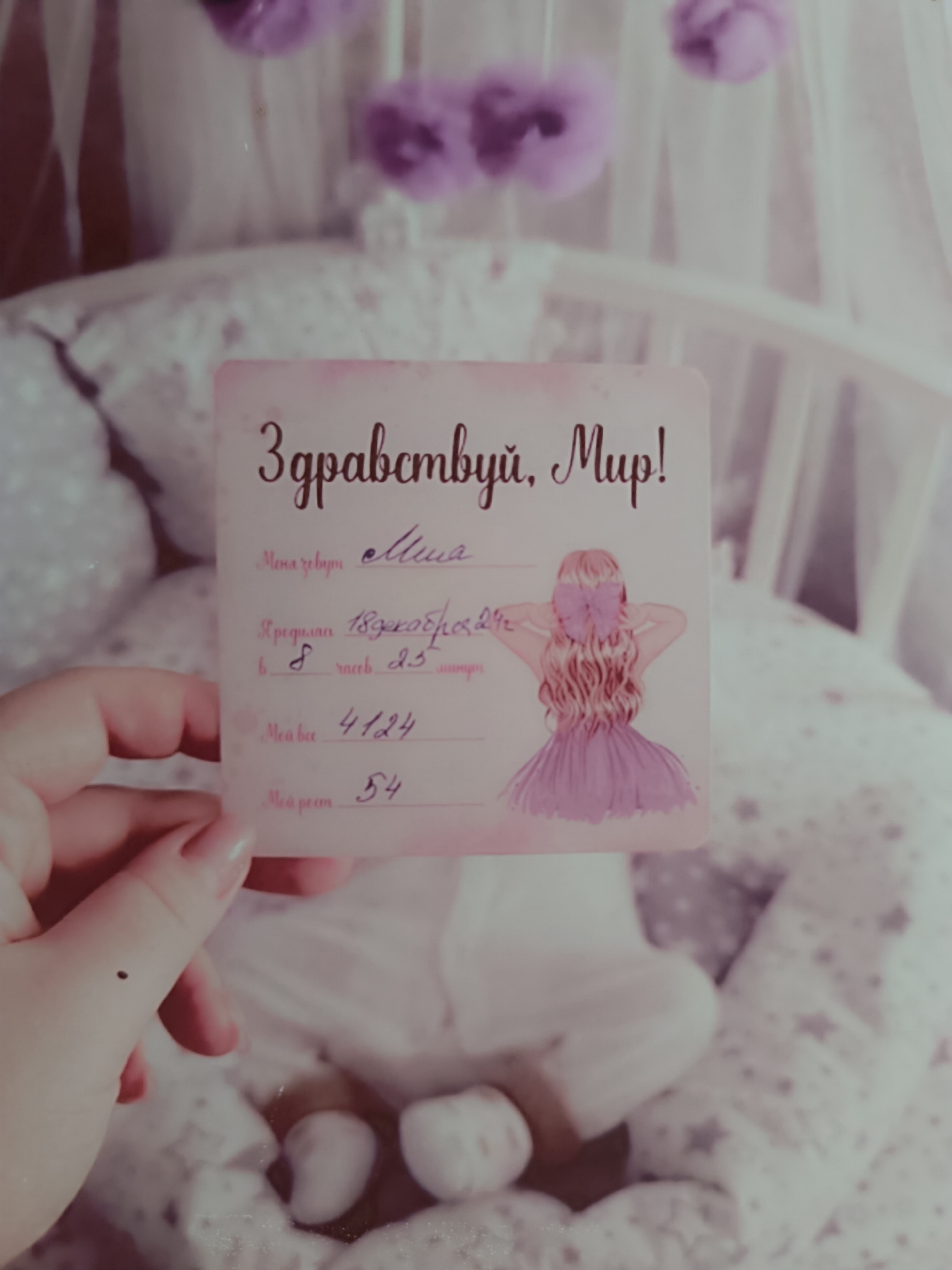 A hand holds up a card that reads "Здравствуй, Мир!" which is Russian for "Hello, World!" The card also includes information such as the name "Miya," the date of birth "18 декабря 29г," the time of birth "8 часов 23," the weight "4124," and the height "54." The card also includes a drawing of a girl with a bow in her hair wearing a pink dress. The background of the image is blurry, but it appears to be a baby's crib. The location is Chaikovsky, Russia.