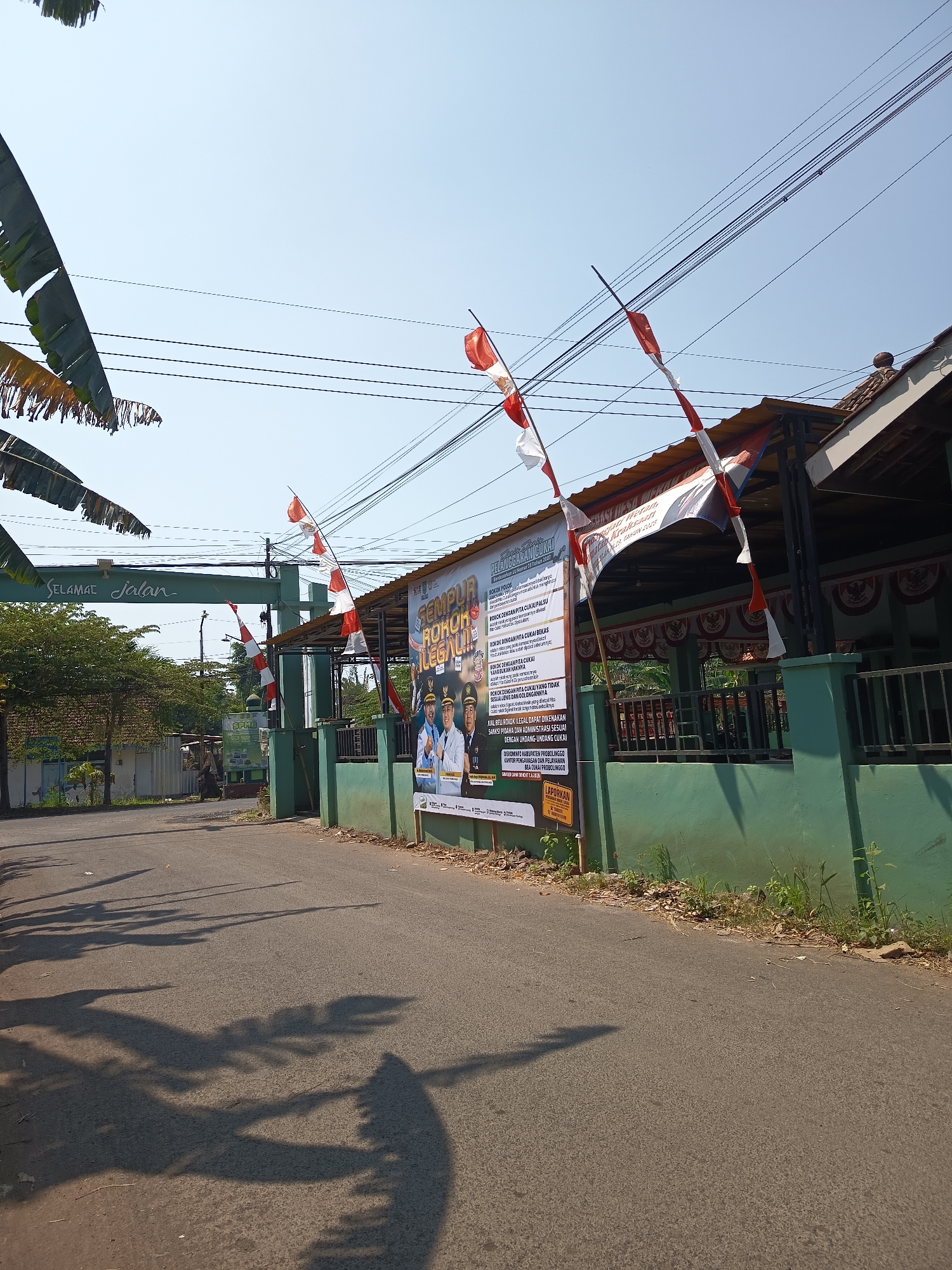 This image depicts an outdoor scene on a sunny day in Kandangjati Wetan, Indonesia. A paved road runs diagonally from the bottom left to the top right, casting long shadows from unseen trees onto its surface. On the right side of the road, a green, low-lying wall runs parallel to the road. Behind the wall, a building with a red-tiled roof is visible, and a dark-colored railing runs along its front. Above the wall and building, a series of electrical wires crisscross the clear blue sky.
Adjacent to the building on the right, a large banner hangs, featuring a central image of two men in formal attire, likely officials, alongside text that appears to be in Indonesian. Several red and white Indonesian flags are mounted on poles, attached to the building and the banner, fluttering in the breeze.
On the left side of the road, a green archway stands over the road with the text "SELAMAT Jalan" (which translates to "Have a good trip" or "Goodbye") in white lettering. Beyond the archway, a cluster of trees and some smaller structures are visible, suggesting a more residential or communal area. Hanging foliage, possibly from banana trees, drapes down from the top left corner of the image. The overall scene suggests a public space adorned for a celebration or event.