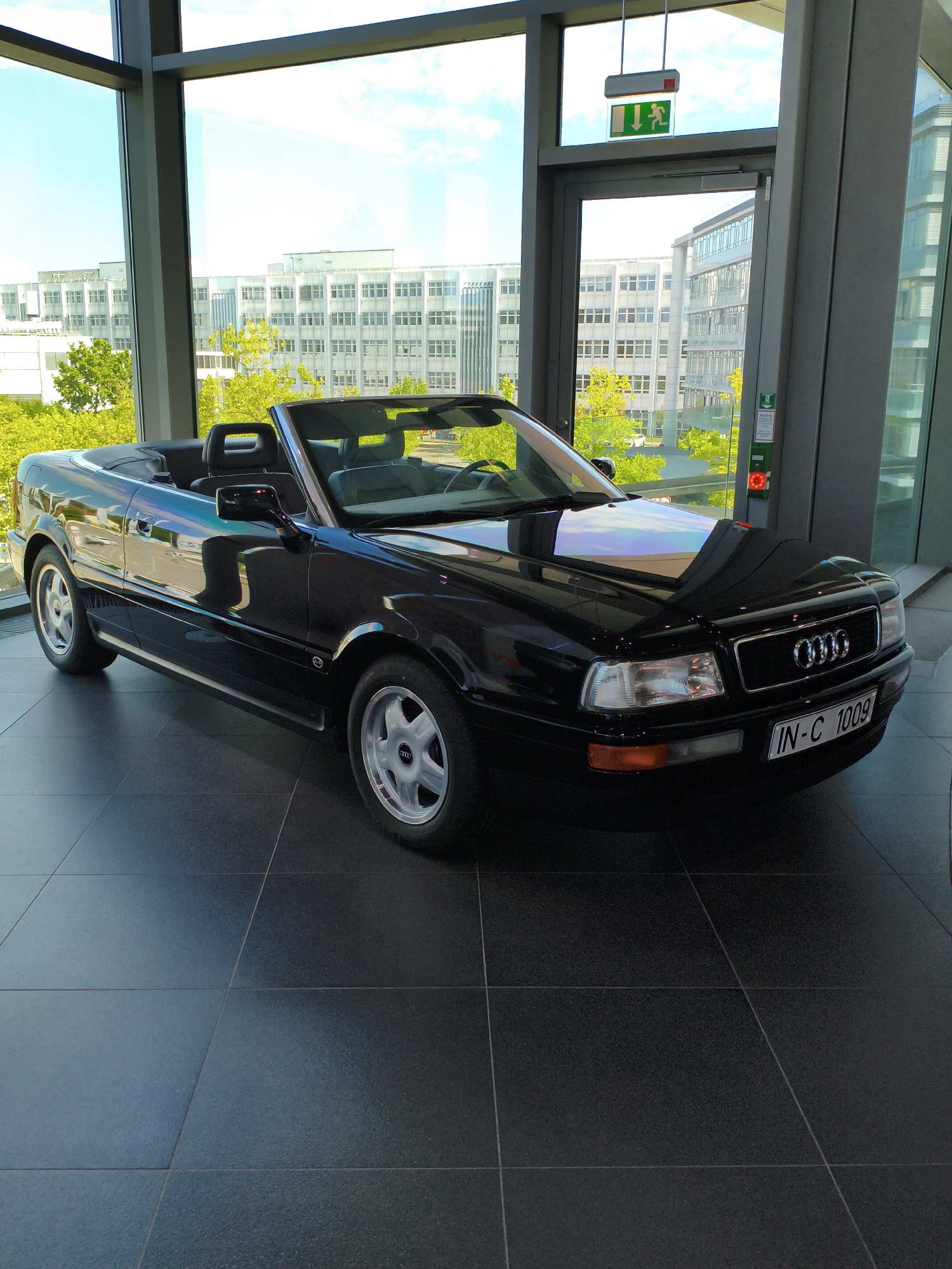 A black Audi convertible is displayed indoors on a dark tiled floor, likely within the Audi Forum in Ingolstadt, Germany. The car is positioned in front of a large window that overlooks a modern cityscape with several large, multi-story buildings and trees. An emergency exit sign with a green running figure is visible above a glass door in the background. The car's license plate reads "IN-C 1009". There are no people present in the image. The lighting is bright, suggesting an indoor showroom environment.