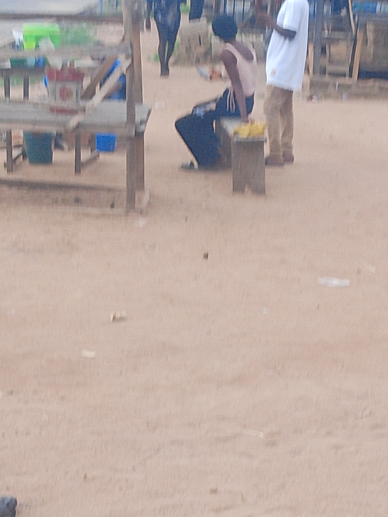 The image depicts an outdoor scene with two individuals. On the left, a person is seated on a wooden bench, wearing dark pants and a light pink top. To their right, a second person stands, dressed in a white t-shirt and tan pants. In front of the seated person, a small wooden structure appears to be holding some yellow items, possibly food. Behind the individuals, there are makeshift structures and tables, suggesting a marketplace or roadside stall. The ground is sandy and appears somewhat wet in places, with a few plastic items scattered around. The lighting suggests it is daytime, but the overall blurriness of the image makes it difficult to ascertain specific details about the time of day or weather. The scene appears to be in Jalingo, Nigeria, based on the provided location context. There is no visible text in the image.