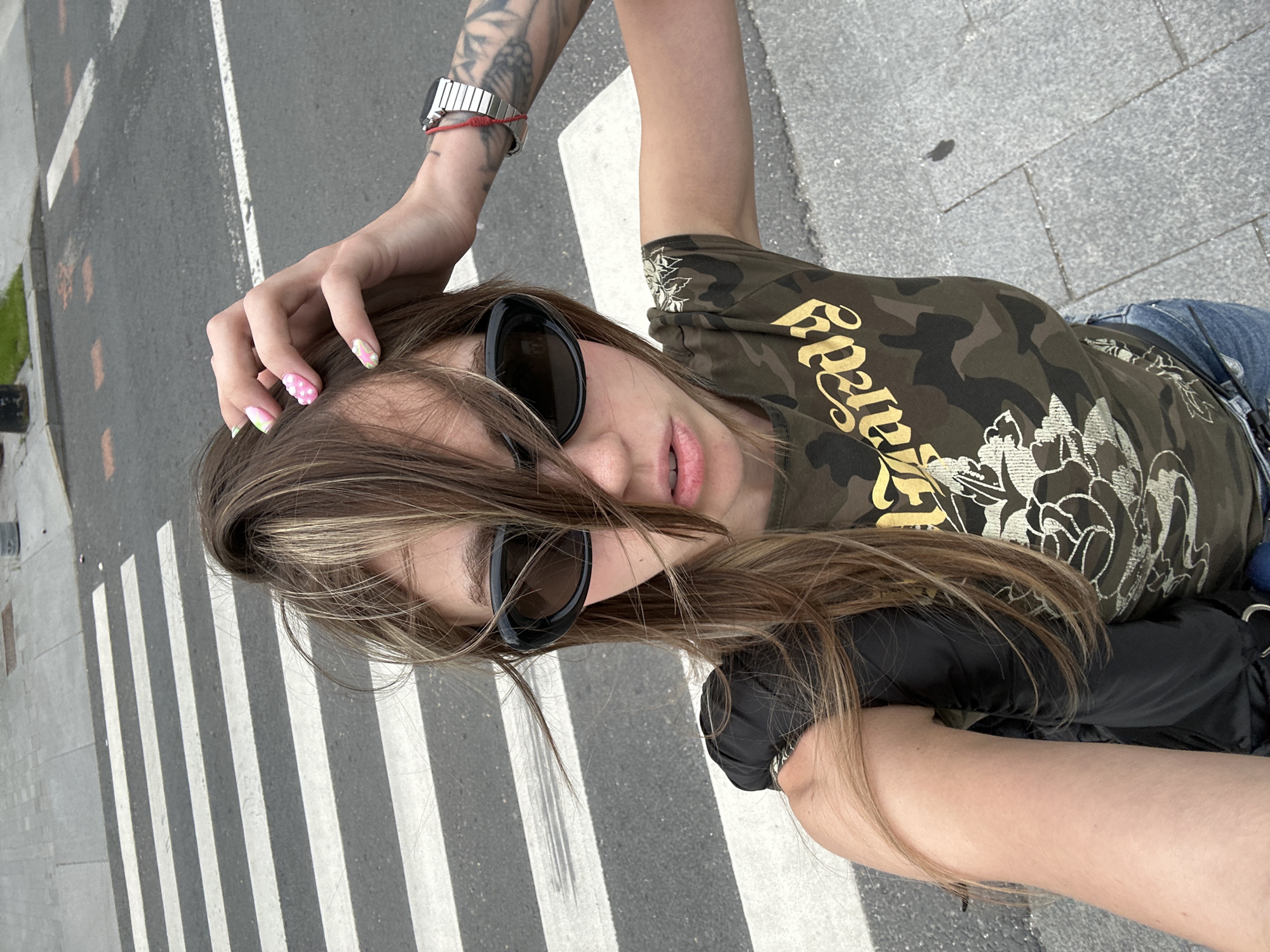 A young woman with light brown hair and bangs covering her eyes is standing on a pedestrian crossing in Moscow, Russia. She is wearing dark sunglasses, a camouflage t-shirt with a gold graphic, and blue jeans. Her right hand is raised, touching her hair, and her left arm is visible, resting by her side. She is also wearing a silver watch with a red string bracelet on her right wrist. The background shows the asphalt road and the white stripes of the pedestrian crossing, indicating an urban environment.