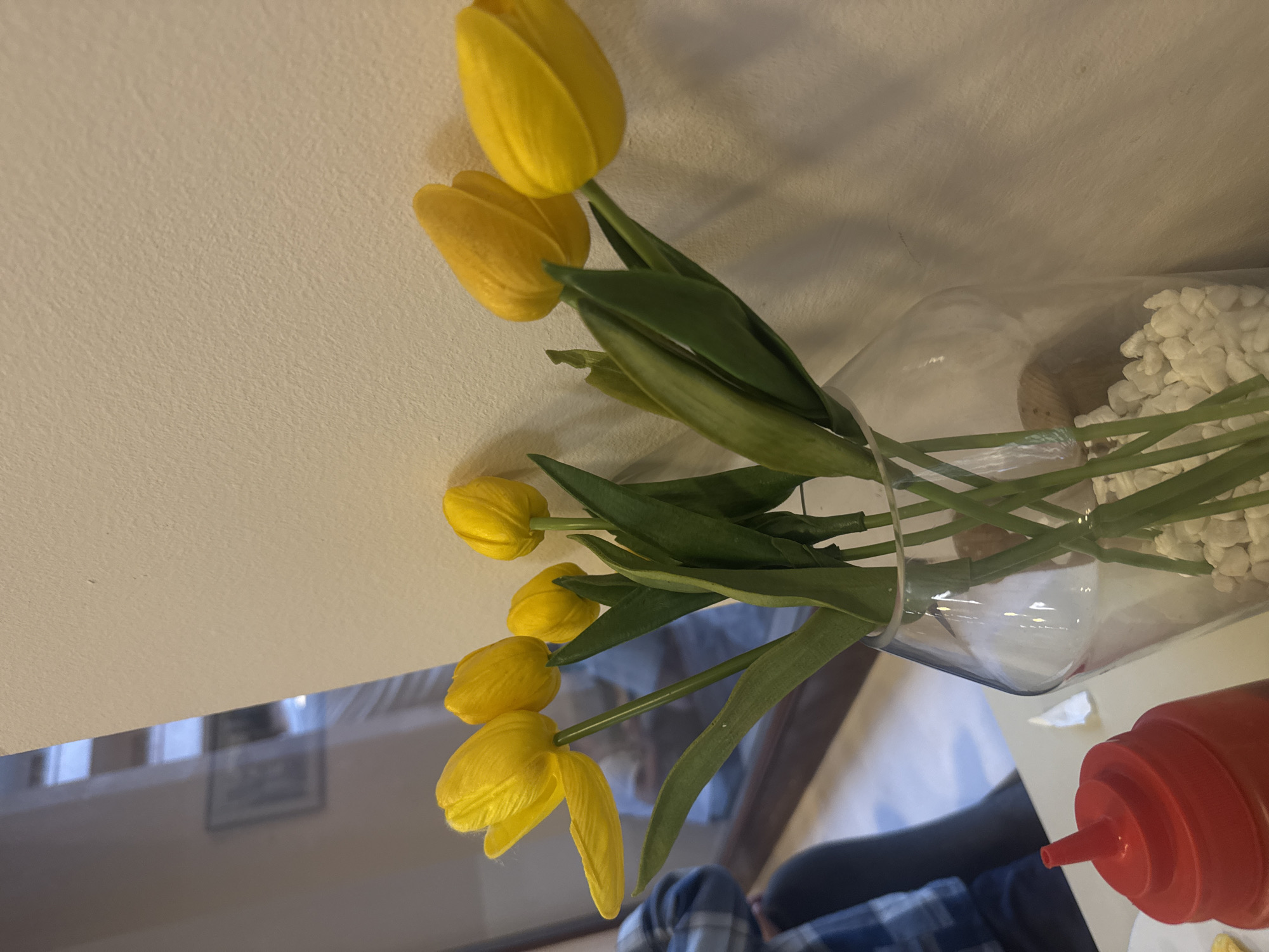 A table top view shows a vase filled with yellow tulips and white decorative rocks, situated in a scene that also includes a red bottle, possibly containing ketchup. A person wearing a blue and white plaid shirt is partially visible. The setting is a restaurant located in Yerevan, Armenia. The lighting is moderate, casting soft shadows.