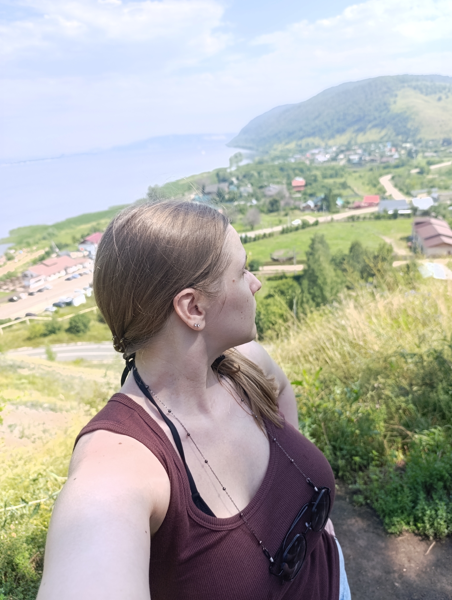 A woman is taking a selfie from a high vantage point overlooking a village and a body of water. She is wearing a brown tank top and a necklace with dark beads. Sunglasses are hanging on her tank top. Her hair is pulled back in a ponytail. The background shows a green, hilly landscape with scattered houses and a road winding through the terrain. In the distance, a large body of water meets a hazy horizon under a partly cloudy sky. The scene is likely set in Solnechnaya Polyana, Russia, known for its scenic views and proximity to the Volga River.