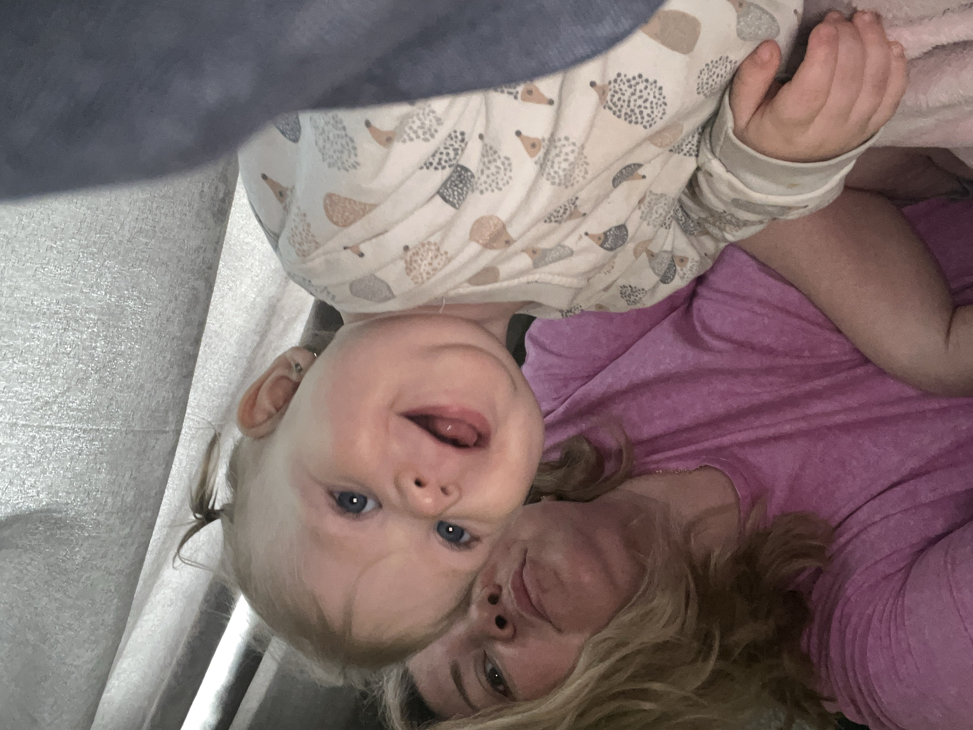 The image, taken in Фоки, Russia, presents a heartwarming, close-up portrait of an adult and a baby lying together, captured from an overhead perspective, resembling a selfie. The original image is rotated, showing the baby positioned above and to the left of the adult when viewed upright.
The baby, with fair skin, bright blue eyes, and blonde hair styled in small pigtails, is looking directly at the camera with a wide, joyful smile, her mouth slightly open with her tongue peeking out. She is wearing a cream-colored long-sleeved top adorned with a repeating pattern of cartoon hedgehogs in various shades of brown, grey, and black. A small ear piercing is visible on her left ear.
Lying closely next to the baby, an adult with blonde hair and fair skin is visible, also looking towards the camera with a gentle smile. She is wearing a solid magenta or purple long-sleeved top. Their heads are close, suggesting a moment of affection and bonding in what appears to be a cozy indoor setting.
The background features muted tones: a dark grey or blue fabric partially covers the upper left, and a lighter grey, textured surface (possibly upholstery or bedding) fills the rest of the left side. A soft pink blanket or fabric is visible near the adult's right arm. The overall atmosphere is intimate and happy, capturing a tender moment between the two individuals. There is no visible text.