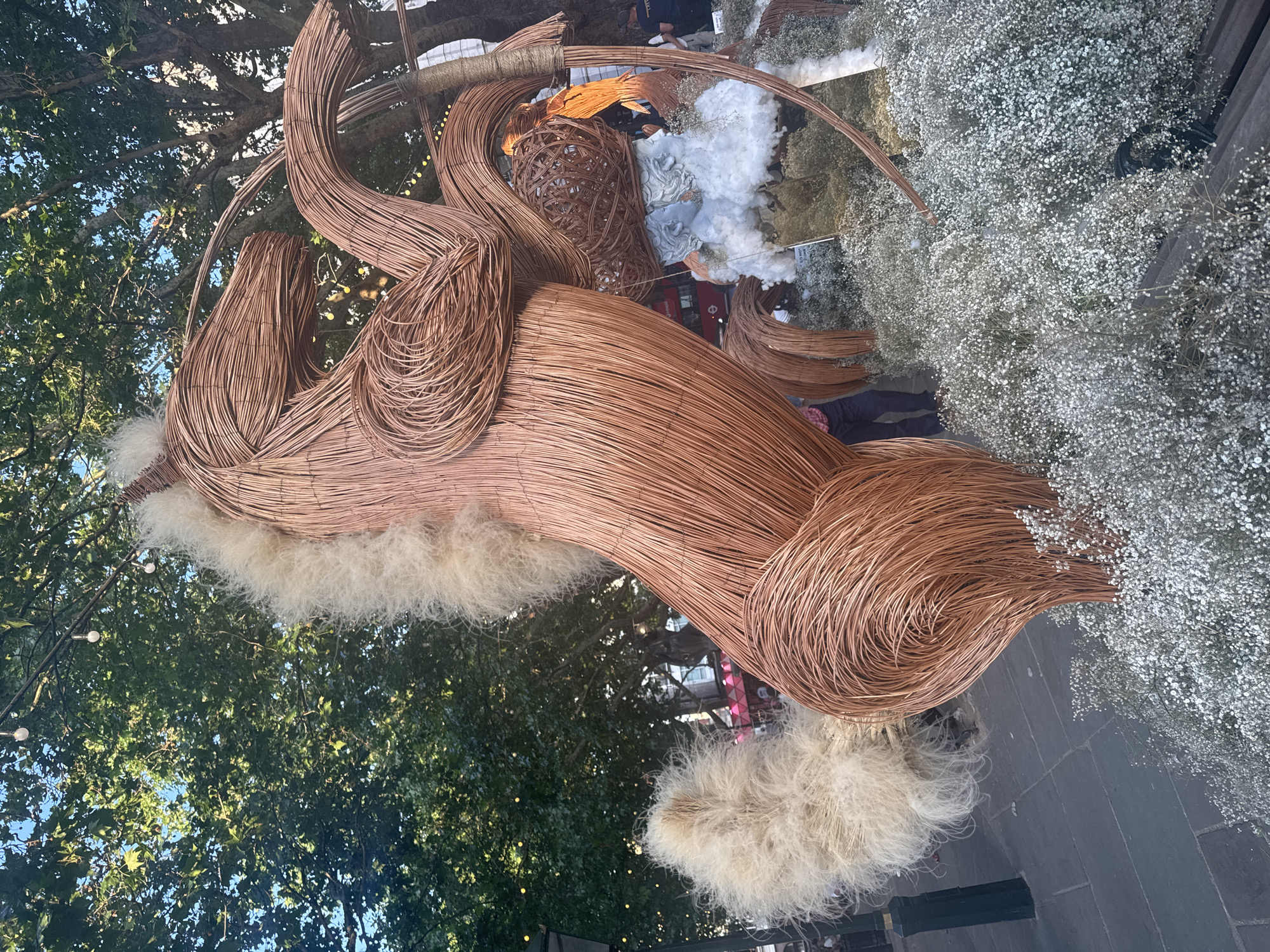 An eye-level, outdoor shot presents a large, brown woven sculpture resembling an animal, adorned with fluffy white material and white flowers. The location is a city, with trees and building facades visible in the background. The sculpture is constructed from woven strands. The fluffy white material is attached at different points. The white flowers form a dense cluster to the right of the sculpture. Below the sculpture, people stand on a stone-paved ground.